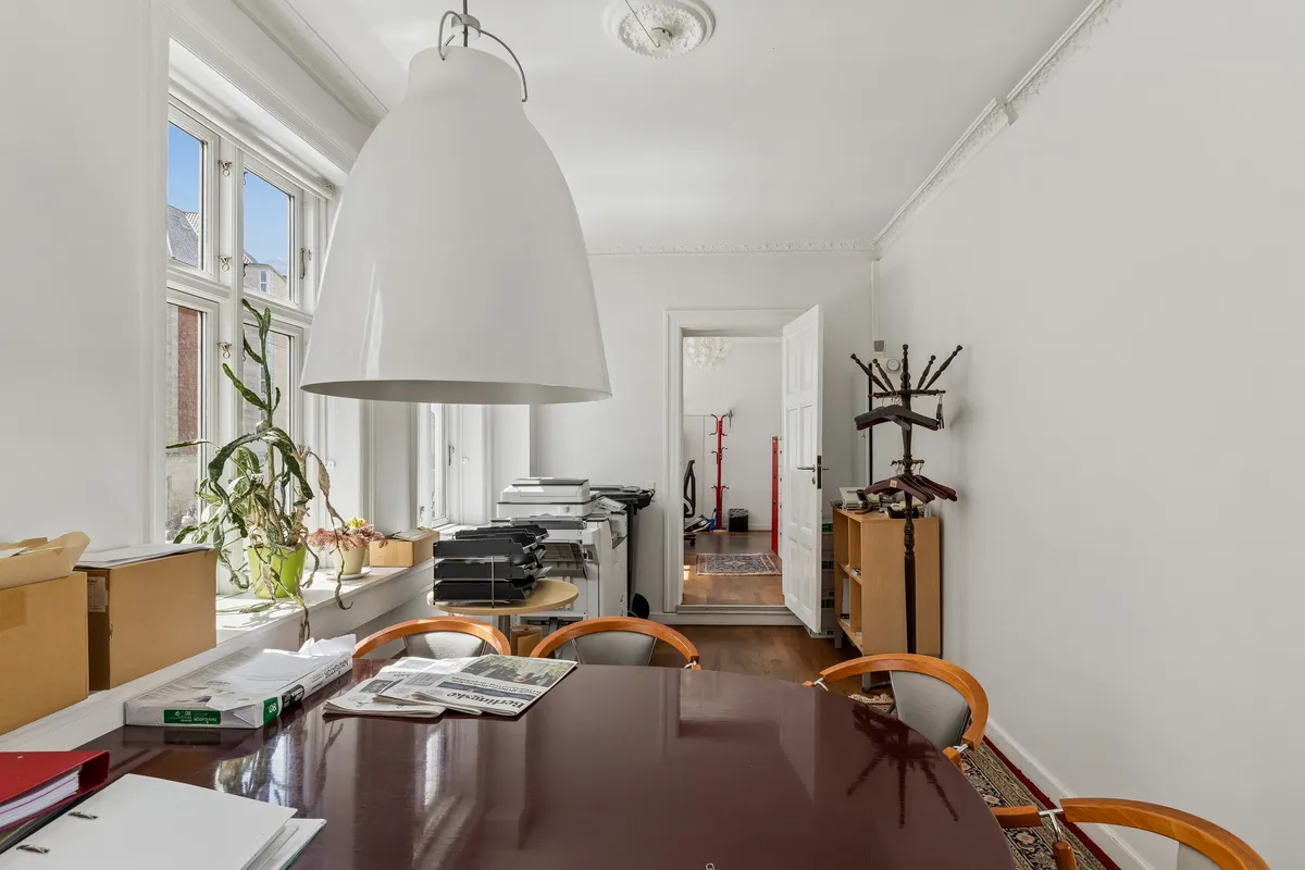 Kontorrum med lyse gulvbelægning og højloft, fuld af møbler og plante. En stor borde med aviser, mapper og en lysekron.