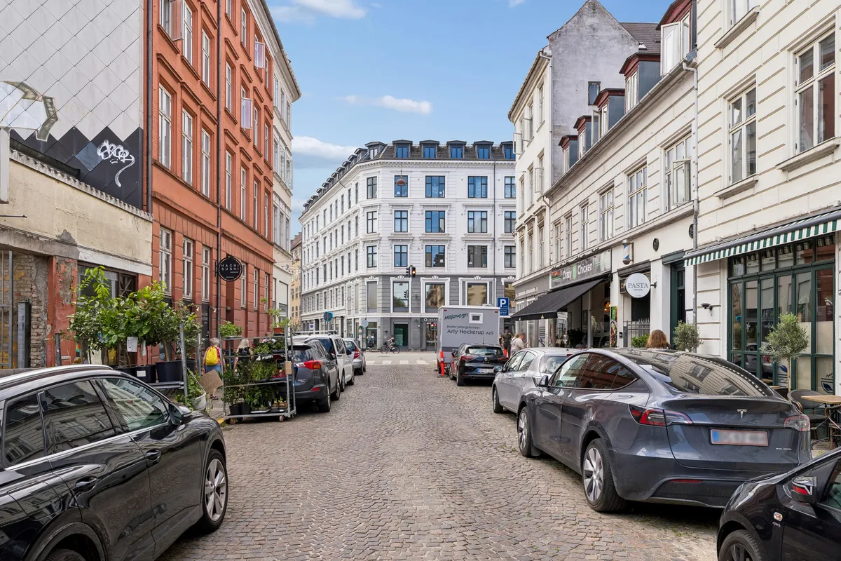En bilparkeret gade med Tesla Model 3 og forskellige butikker, hvoraf én er Pasta Man og en anden har en grøn og hvid bue.
