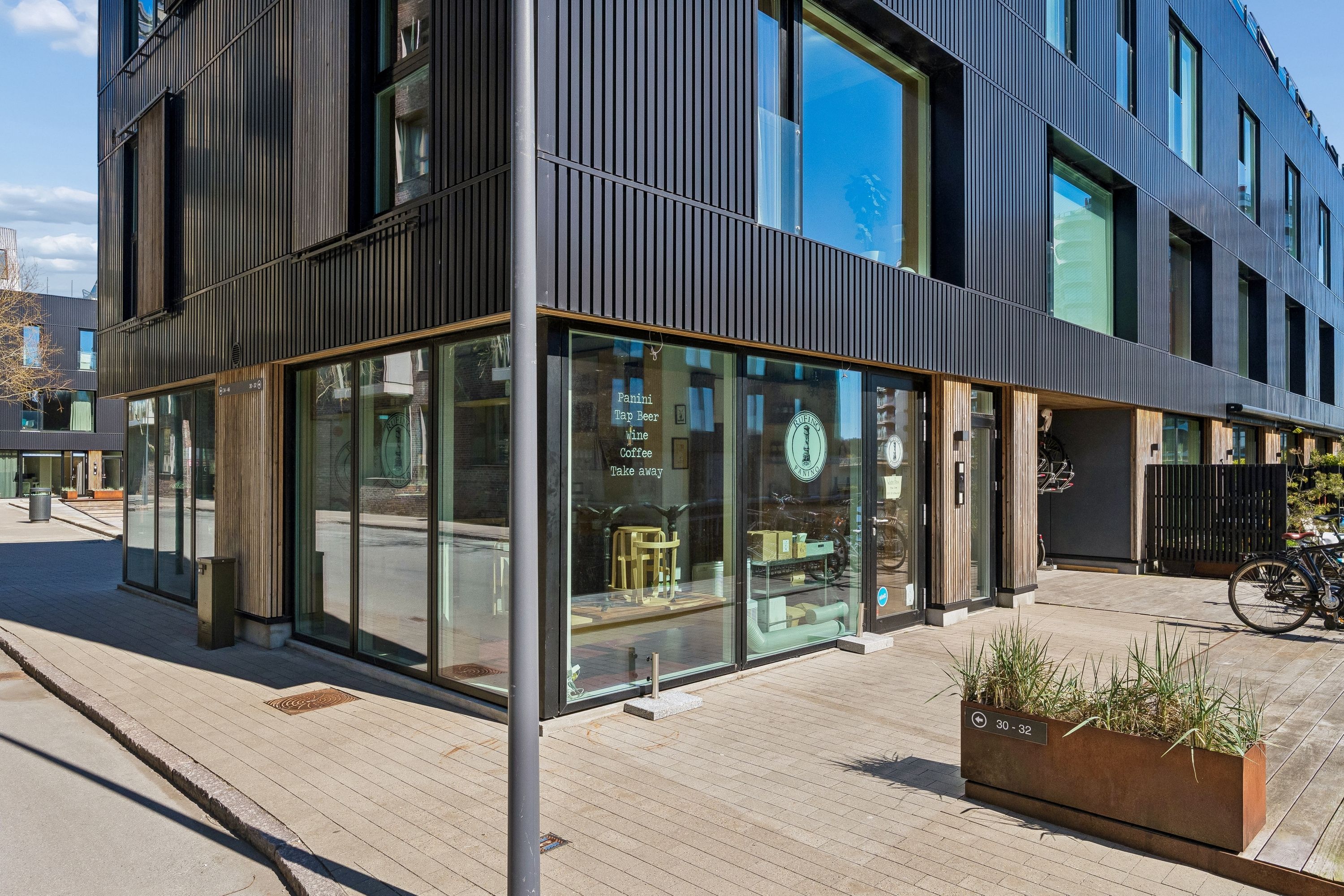 Glass facade og vinduer på butik. Plante i stålpotte på fortov. Cykler parkeret udenfor. Vinduesskilte med teksten Panini Tap Beer Wine Coffee Take away.