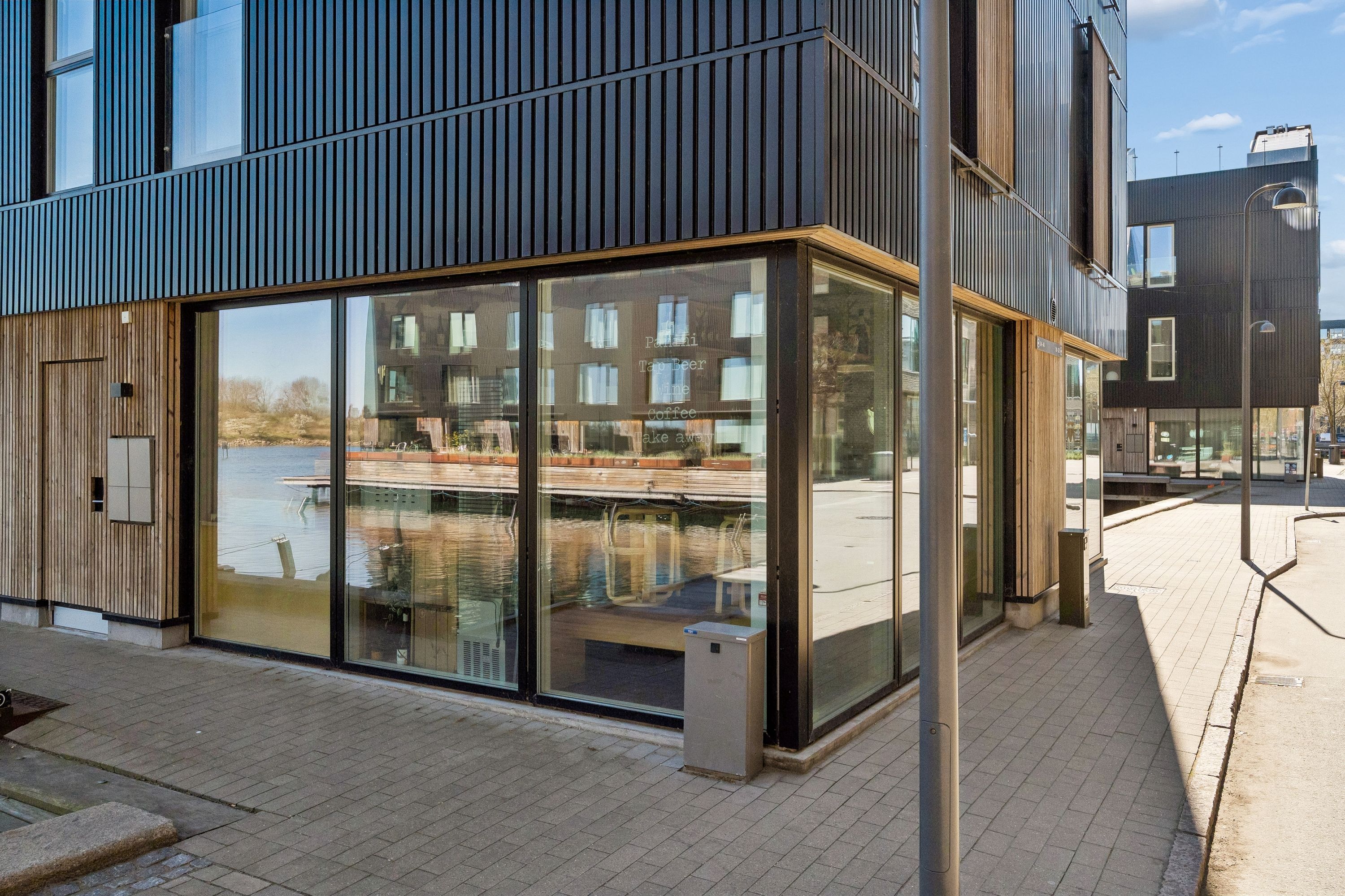 Moderne bygning med glashuse og træindslag. En butik med skilte, der viser tappøl og kaffe. En sø med både og planter i baggrunden. En møbelbutik med udsalgsartikler på terrassen