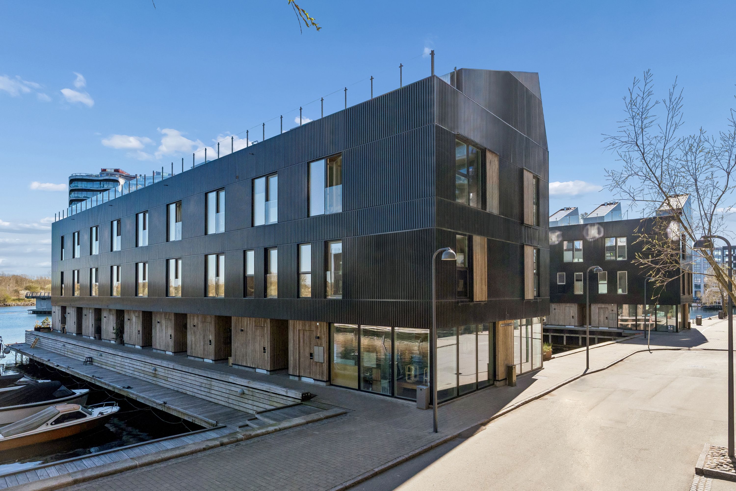 Moderne bygning med glasvinduer og mørk facade ligger ved vandet, med parkerede både og gadebelys langs siden