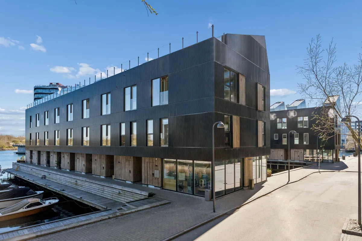 Moderne bygning med glasvinduer og mørk facade ligger ved vandet, med parkerede både og gadebelys langs siden