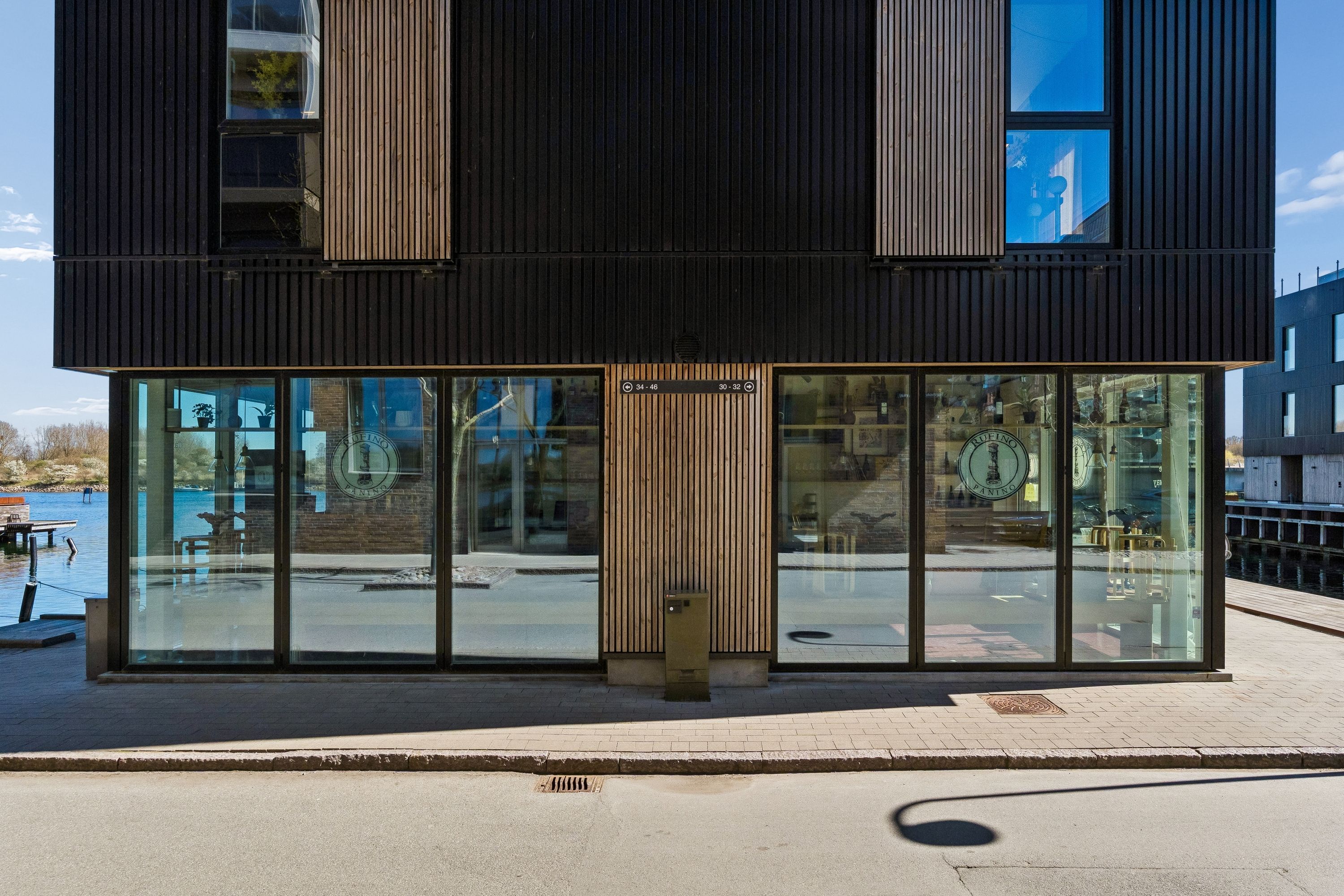 Moderne bygning med glasfacade og mørk beklædning står på kajen. Udenfor er der et skur og en briklægget gang. Bag glasskærmen er der møbler og vægdekorationer.