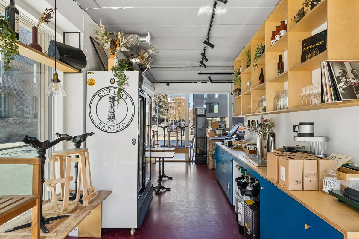 Butik med lyse hjørner, hylder med flasker og grønt. Køleskab med logoet Rufio Panino og gulvbord med stole. Glasvinduer med bygninger og træer udenfor