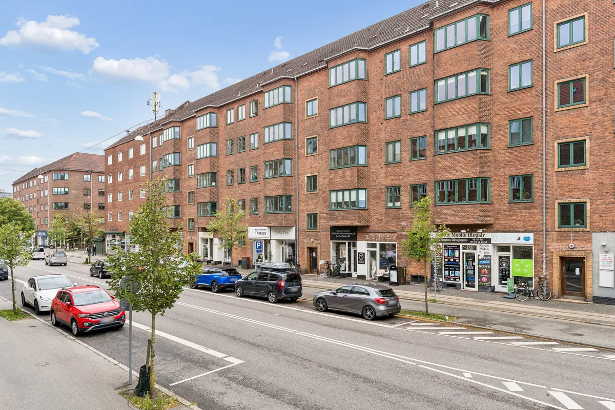 Moderne bygning med butikker og parkerede biler på Borups Alle i Frederiksberg, med smukke træer og grønne ruder