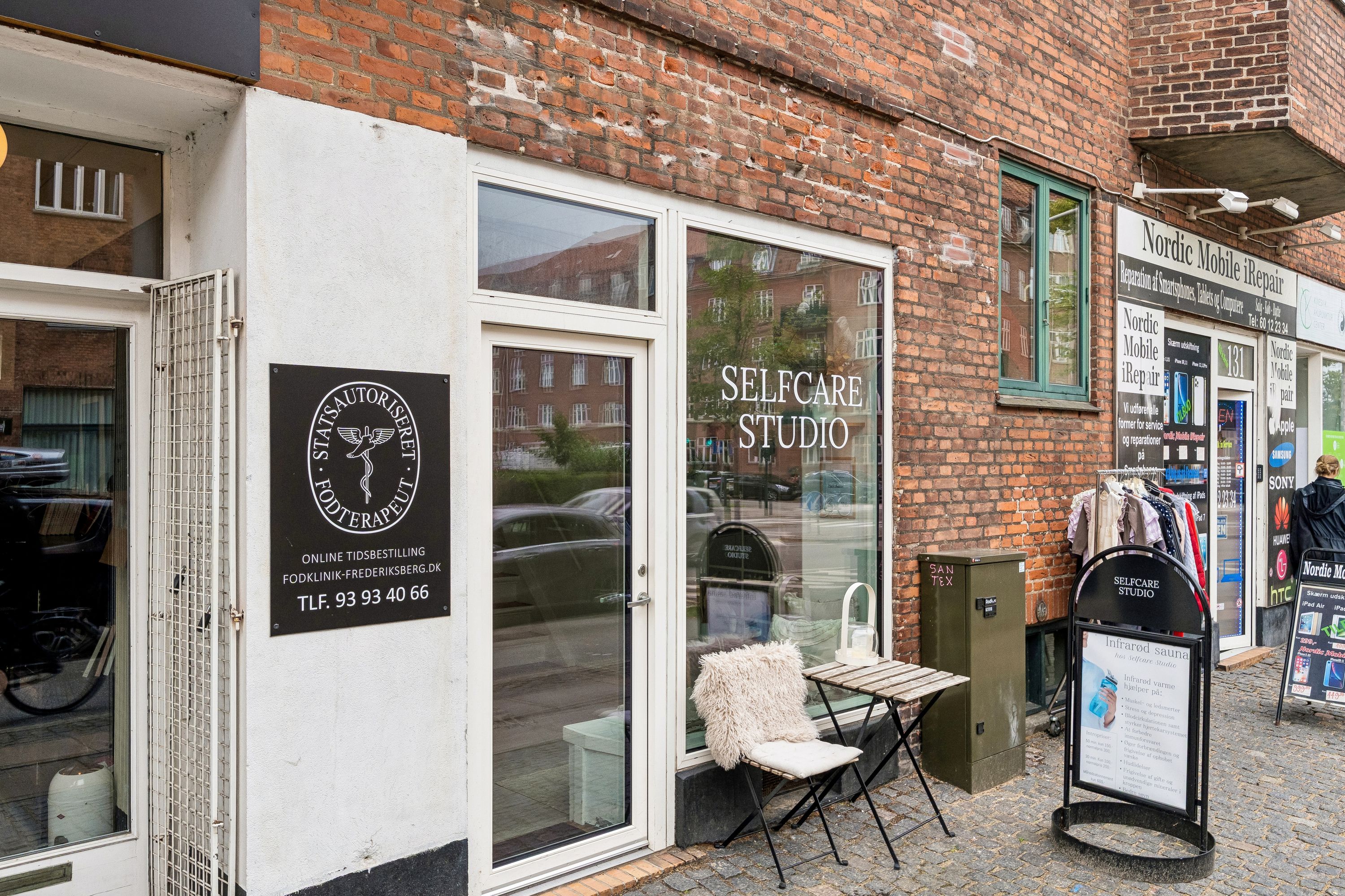 Butiksfront med et vindue og en dør med inskriptioner. En træbænk med hvid pude og et bord er placeret udenfor. Et skilt læser "Selfcare Studio" og et andet skilt med teksten "Infrarød sauna" er placeret på gågaden.