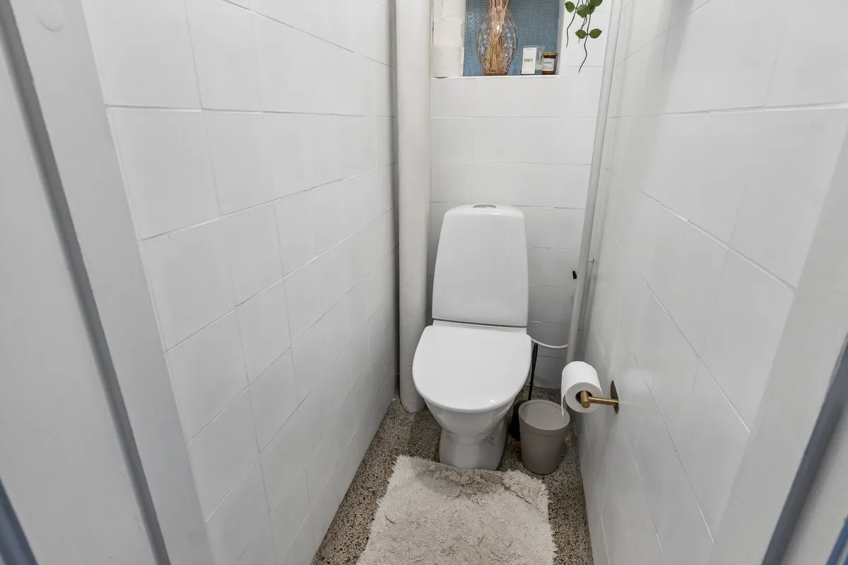 Toilet med hvid toiletbrille, toiletpapir og toiletpapirholder. Væggene og gulvet er hvide og renholdte. Bag toilettet er et lille skab med en vase og andre genstande.