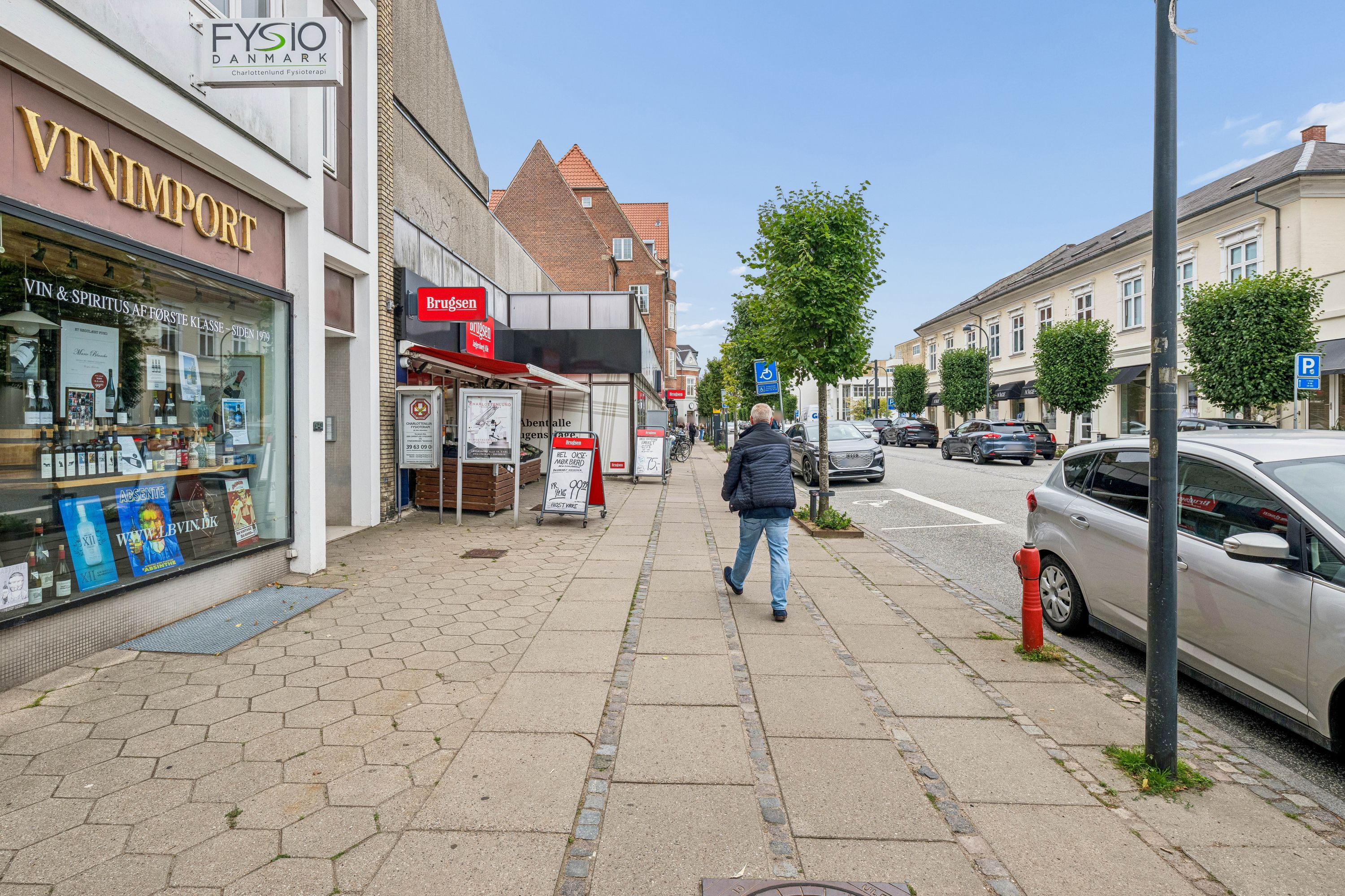 En mand går på gågaden i Charlottenlund med biler og parkerede biler i baggrunden. Foran ham er der flere forretninger og butikker, herunder en vinbutik og en frisør.