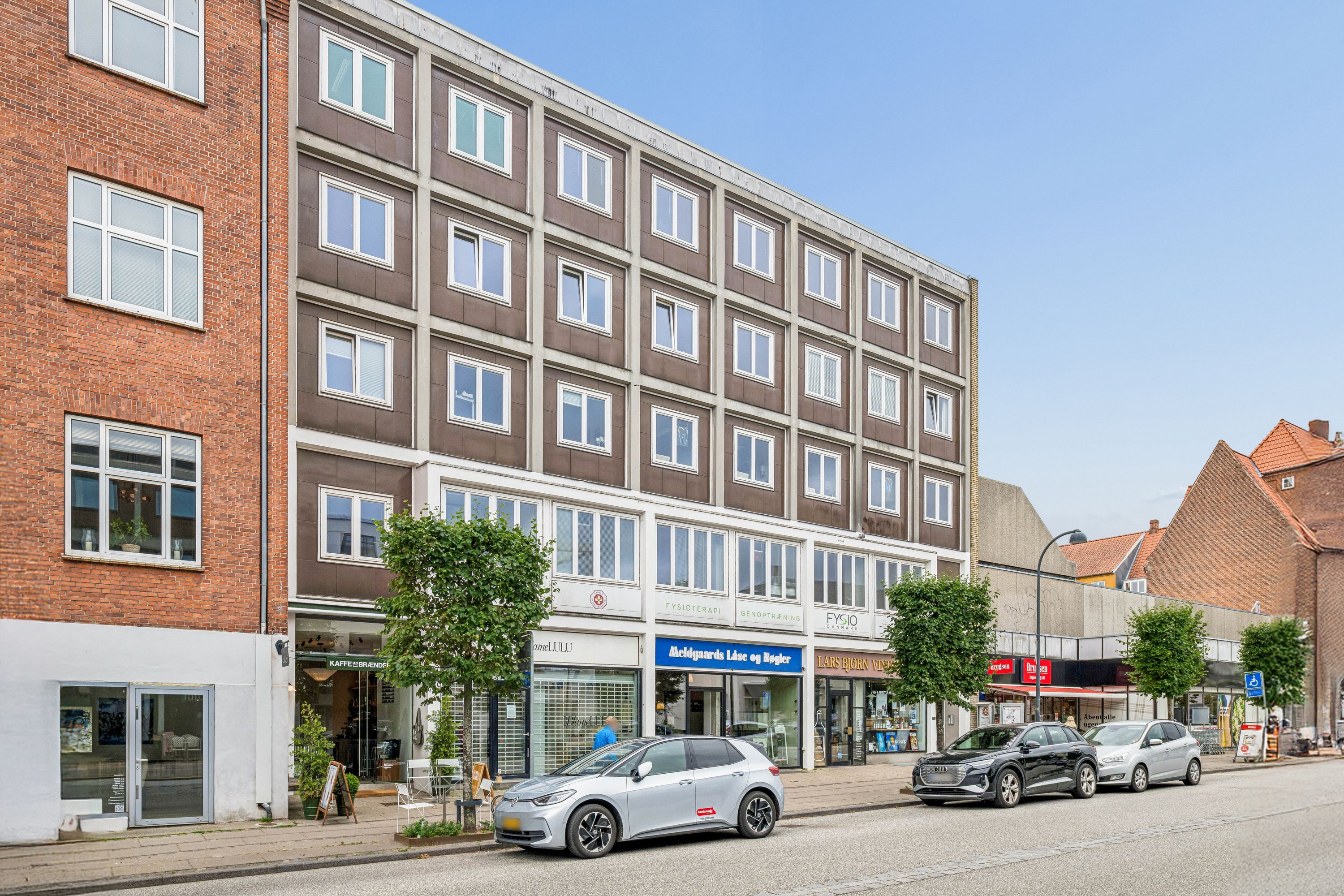 På Jægersborg Alle i Charlottenlund står to biler parkeret. Der er to træer og et bord med stole. Der er flere lokaler med forretninger, hvoraf en er en café.