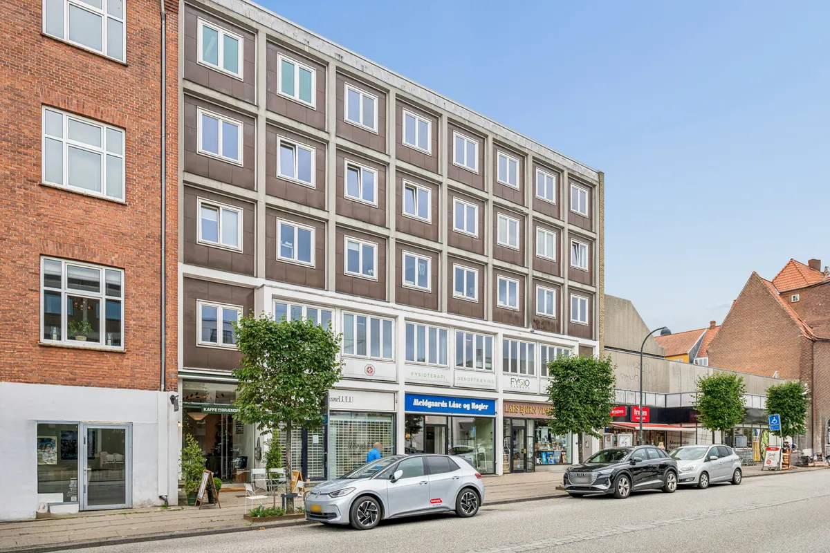 På Jægersborg Alle i Charlottenlund står to biler parkeret. Der er to træer og et bord med stole. Der er flere lokaler med forretninger, hvoraf en er en café.