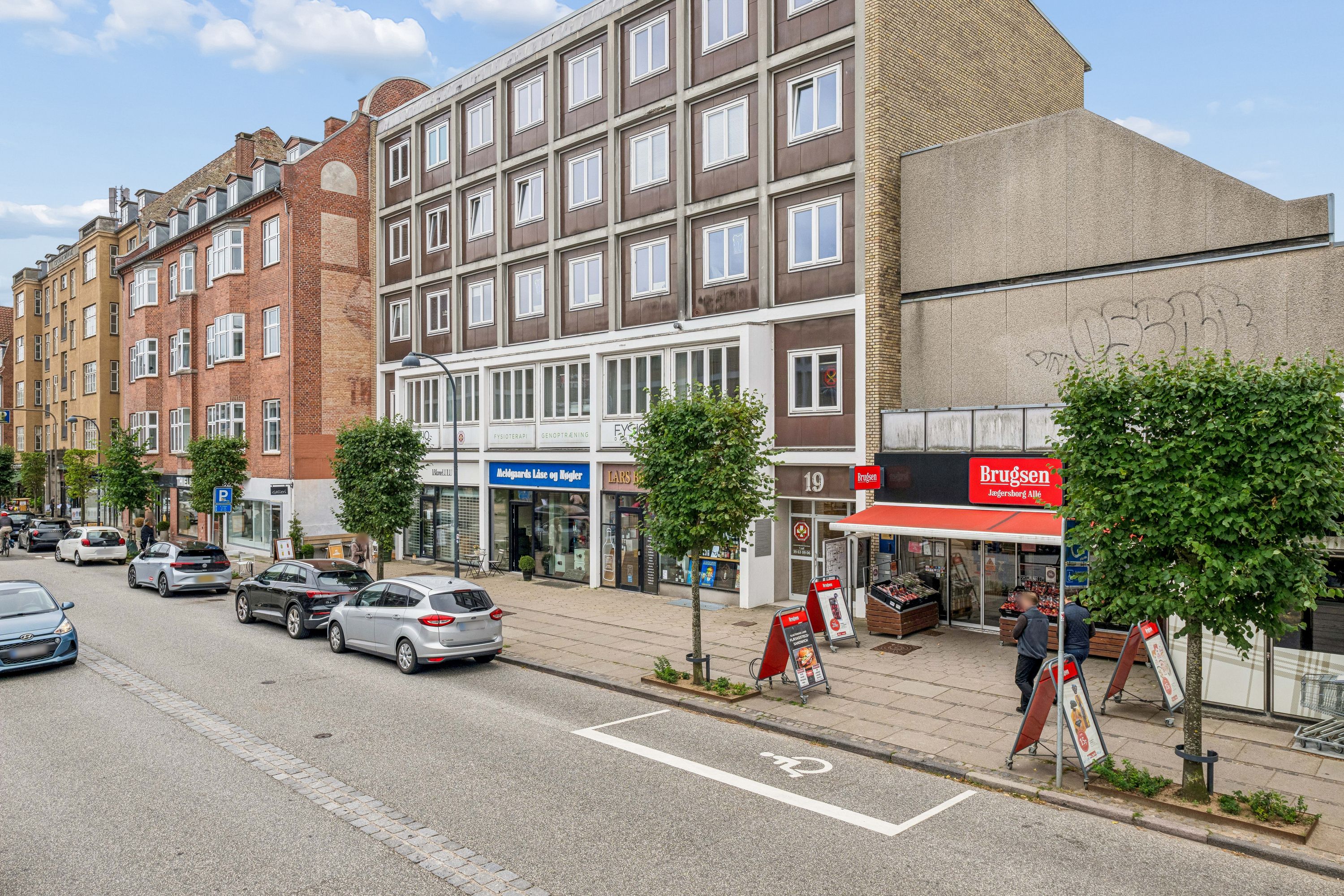 En bygade med parkerede biler, gågade med forretninger og søjler med parkerede cykler. Købmandsforretningen Brugsen er synlig med mennesker der går og taler