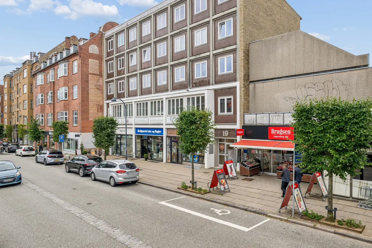 En bygade med parkerede biler, gågade med forretninger og søjler med parkerede cykler. Købmandsforretningen Brugsen er synlig med mennesker der går og taler