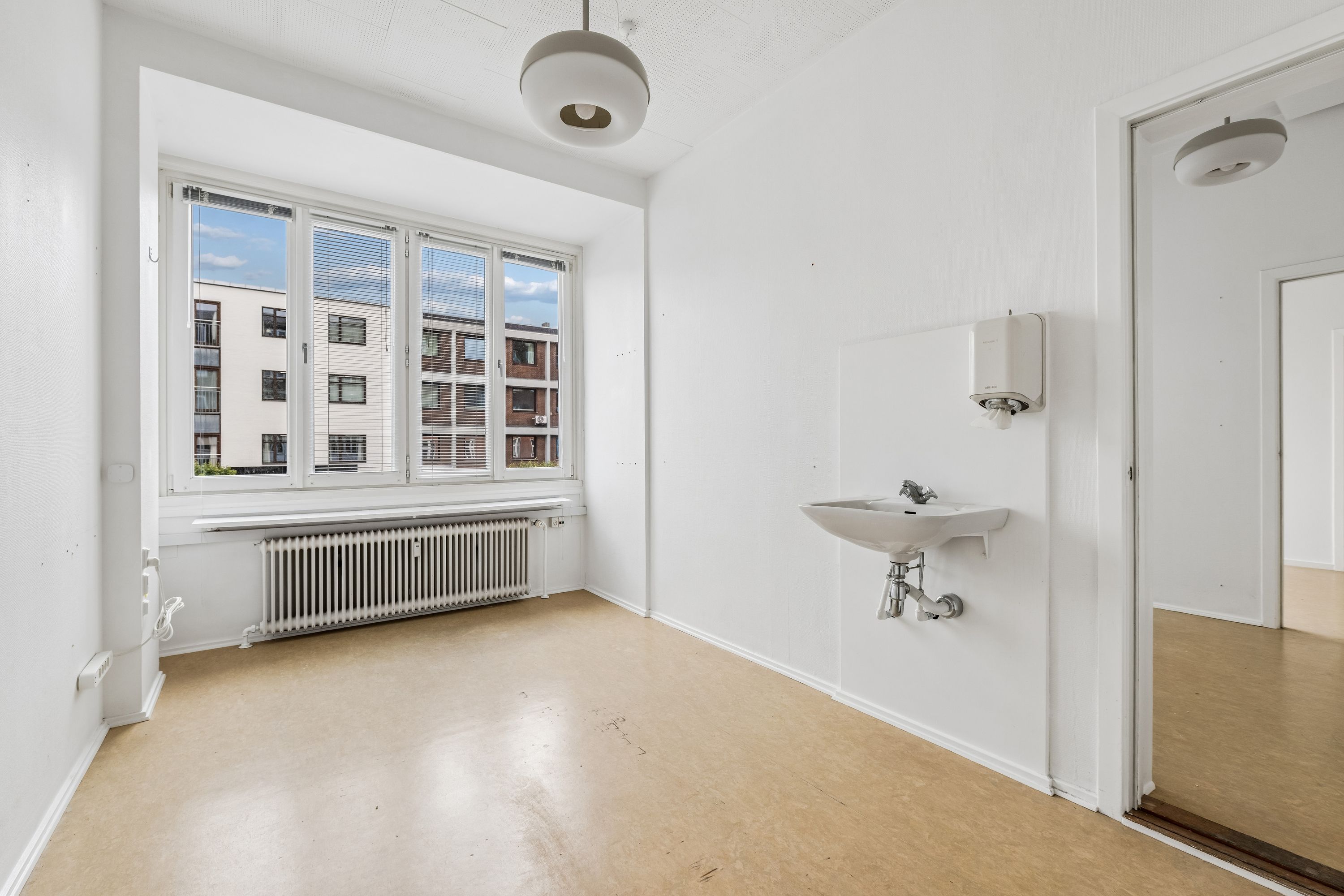 Lejlighed med store vinduer og radiator under vinduet, hvidt toilet med håndklædeautomat, to lyskroner og åben dør