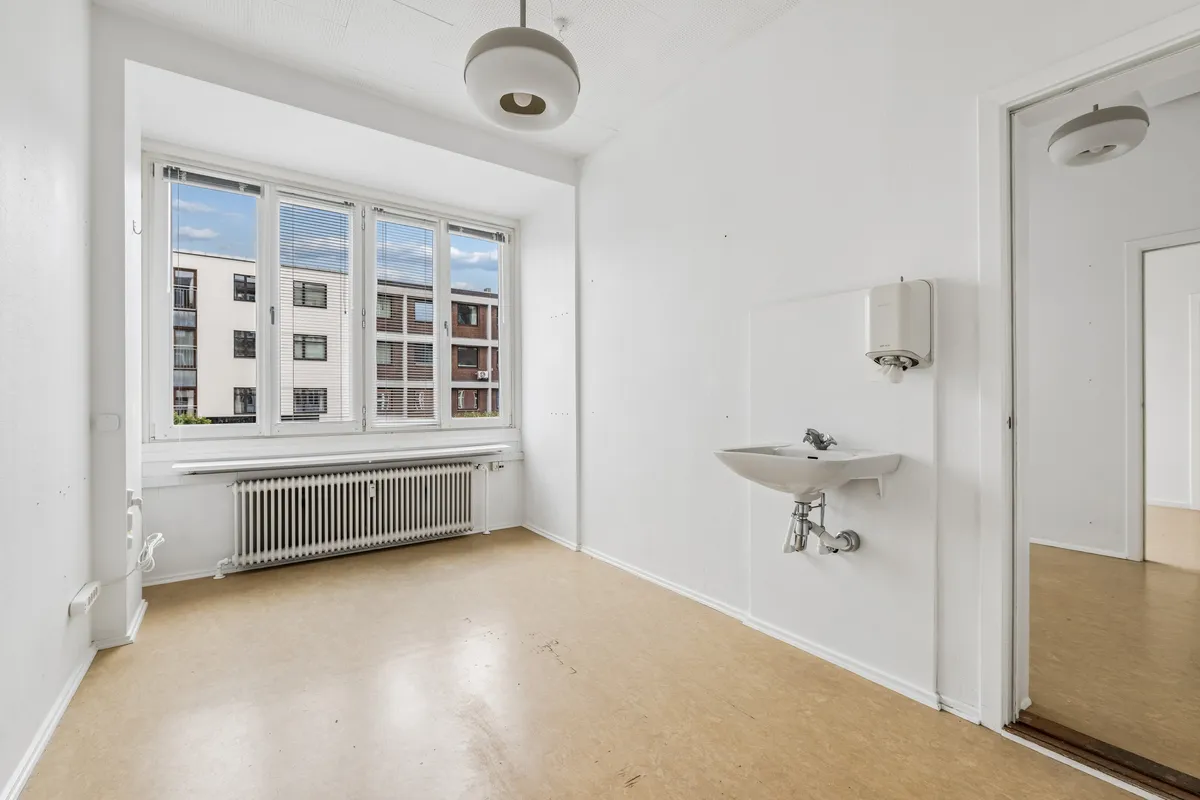Lejlighed med store vinduer og radiator under vinduet, hvidt toilet med håndklædeautomat, to lyskroner og åben dør