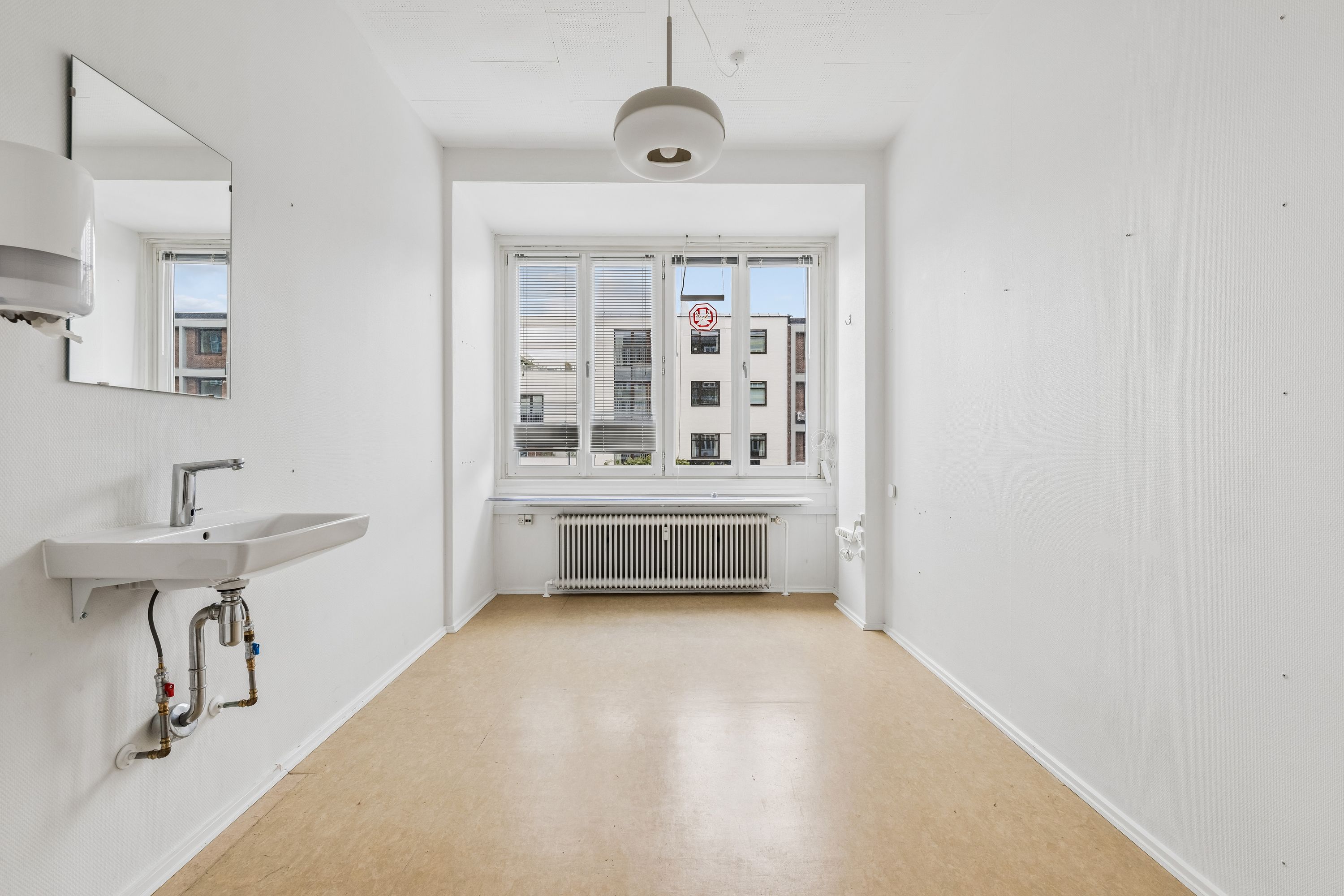 En hvid badeværelsesrum med radiator, vandhaner og spejl samt vinduer med gardiner