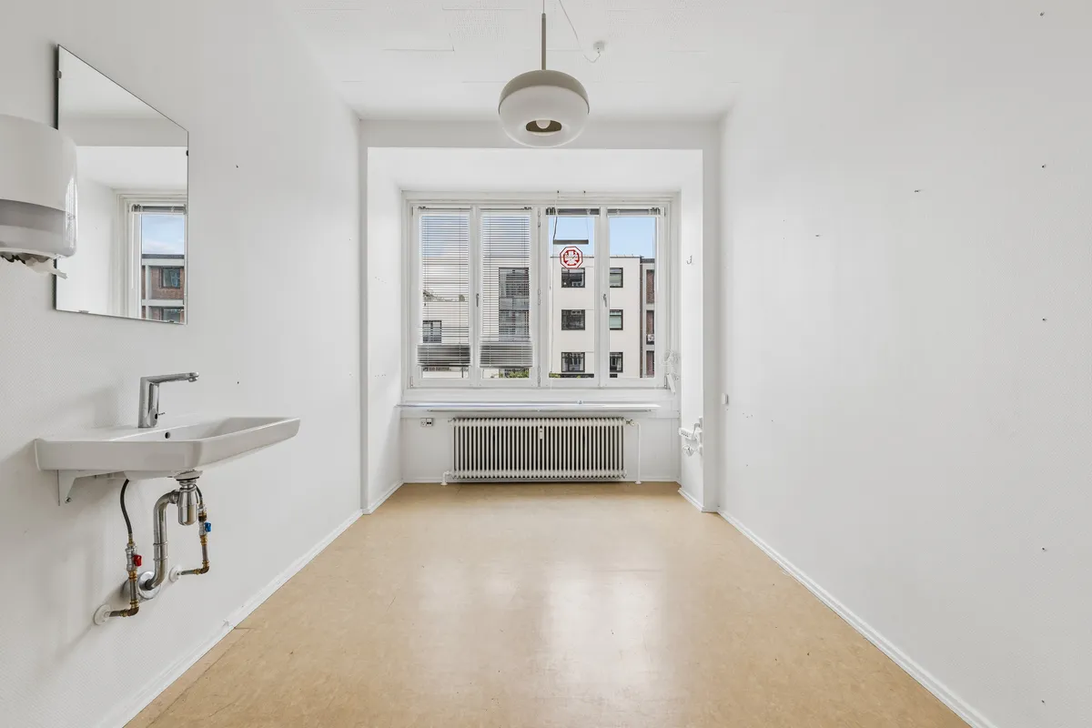 En hvid badeværelsesrum med radiator, vandhaner og spejl samt vinduer med gardiner