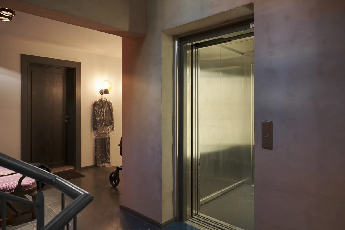 Her ses et glaskledet elevator med åben dør og gulv i gult marmor. Der er skråtag og lysekroner i loftet