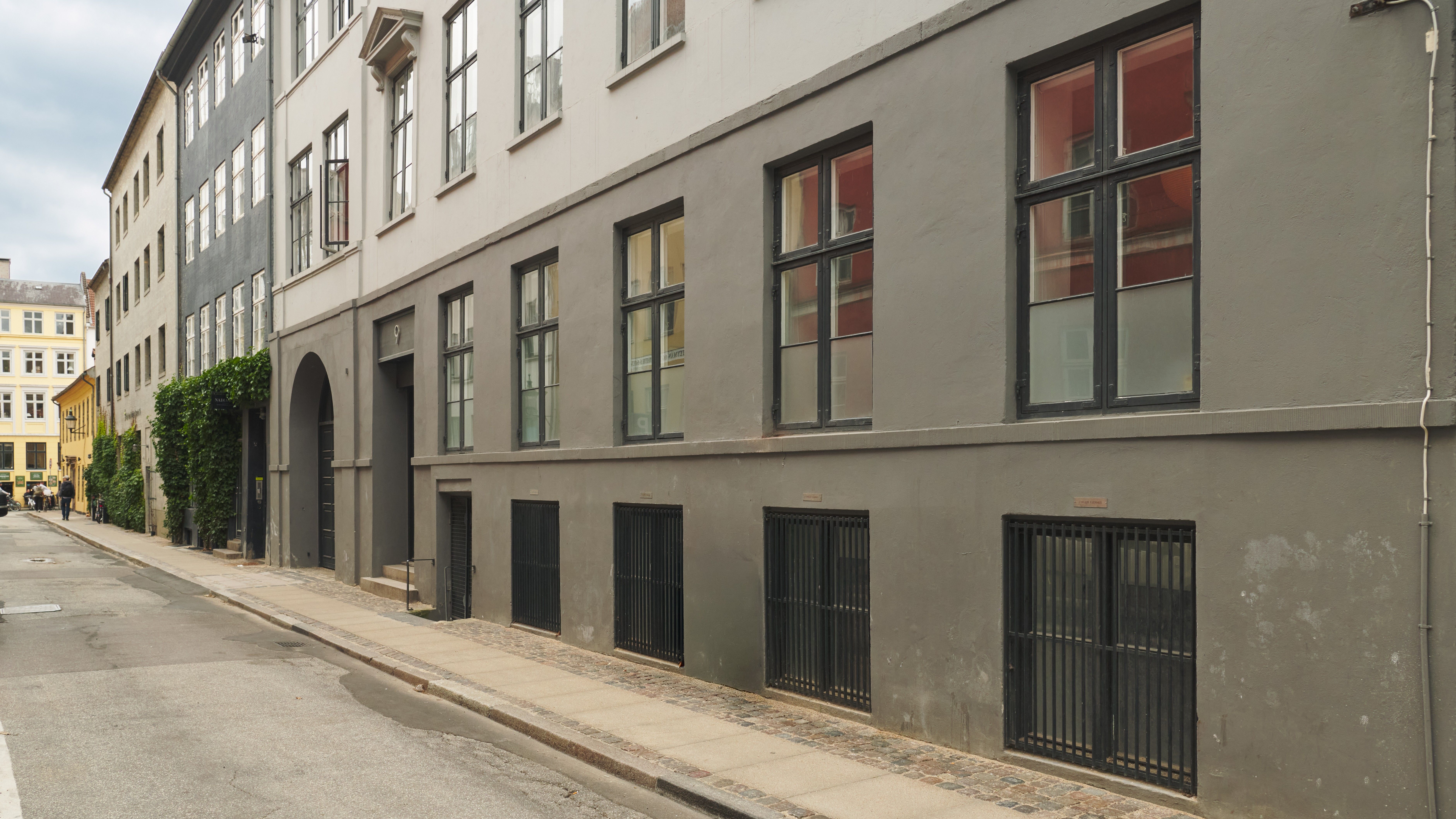 Vejbred med to rækker store, mørke vinduer og høje, lyse stenblokke på bakke. Stærke røde og gule bygninger i baggrunden med grønne planter på facaden.