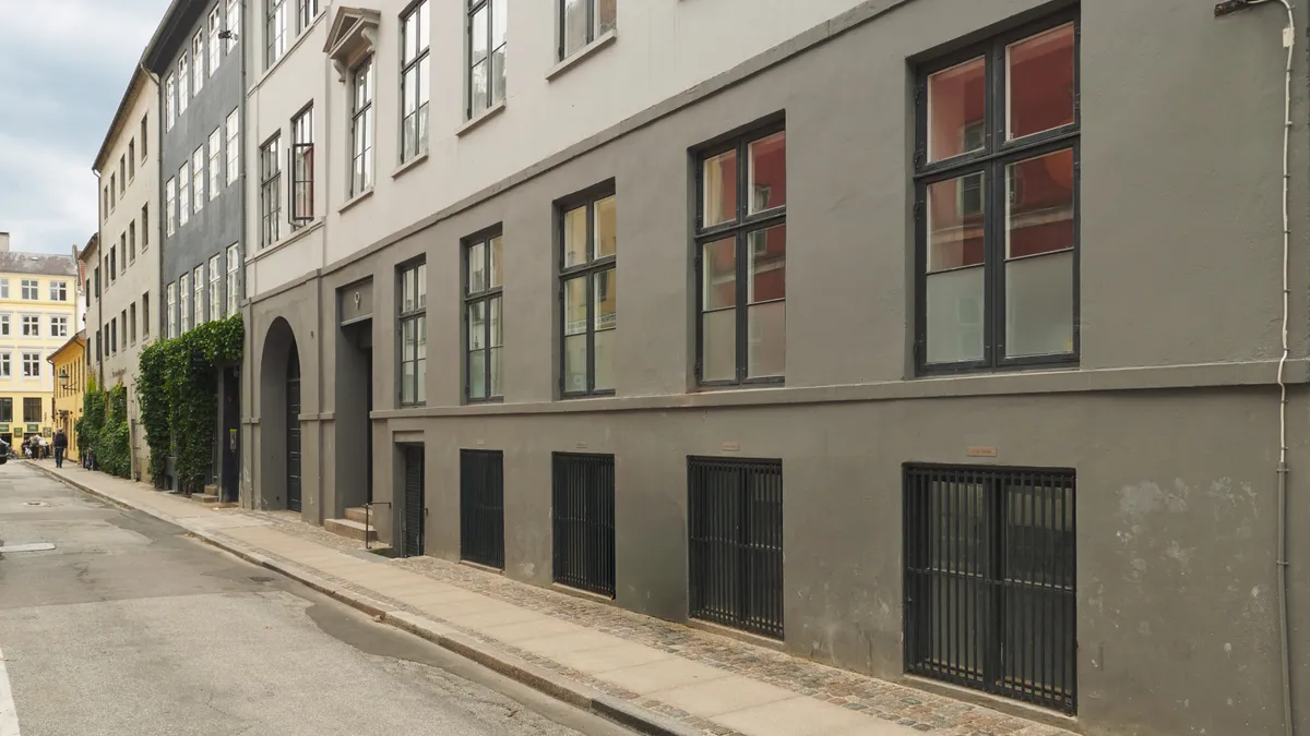Vejbred med to rækker store, mørke vinduer og høje, lyse stenblokke på bakke. Stærke røde og gule bygninger i baggrunden med grønne planter på facaden.