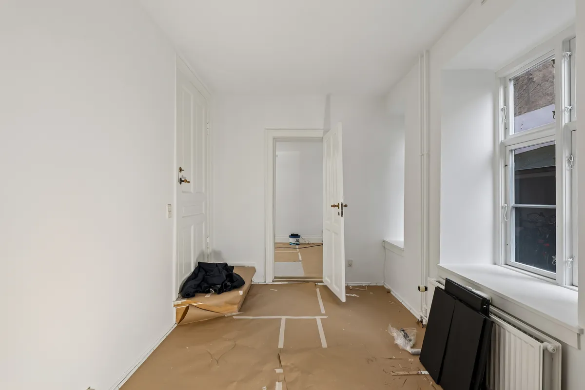 En hvid og tom stue med åbne døre og en række hvide linjer på gulvet og væggen. Et sæt møbler og en radiator står i hjørnet, og vinduet er åbent