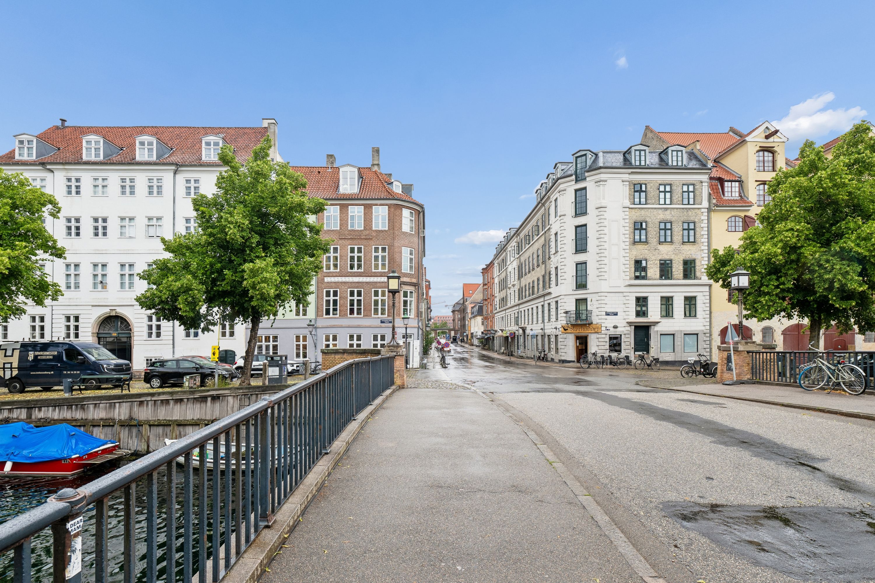 Lejlighed med 80 m² på Sankt Annæ Gade, Christianshavn. Bygning med mange vinduer og buskplanter. Ledig vej med cykler og parkerede biler. Vand med en rød båd og en båd med blåt tæppe.