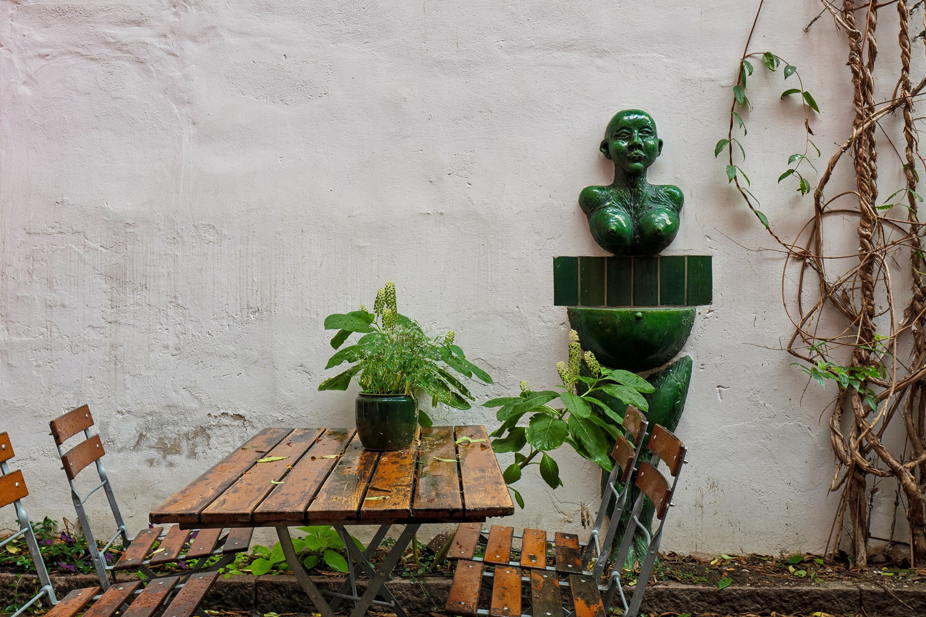 Foto af udendørs bord og to stole med en grøn plante og statue i baggrunden. På bordet ligger et glas og en grøn vase med en plante.
