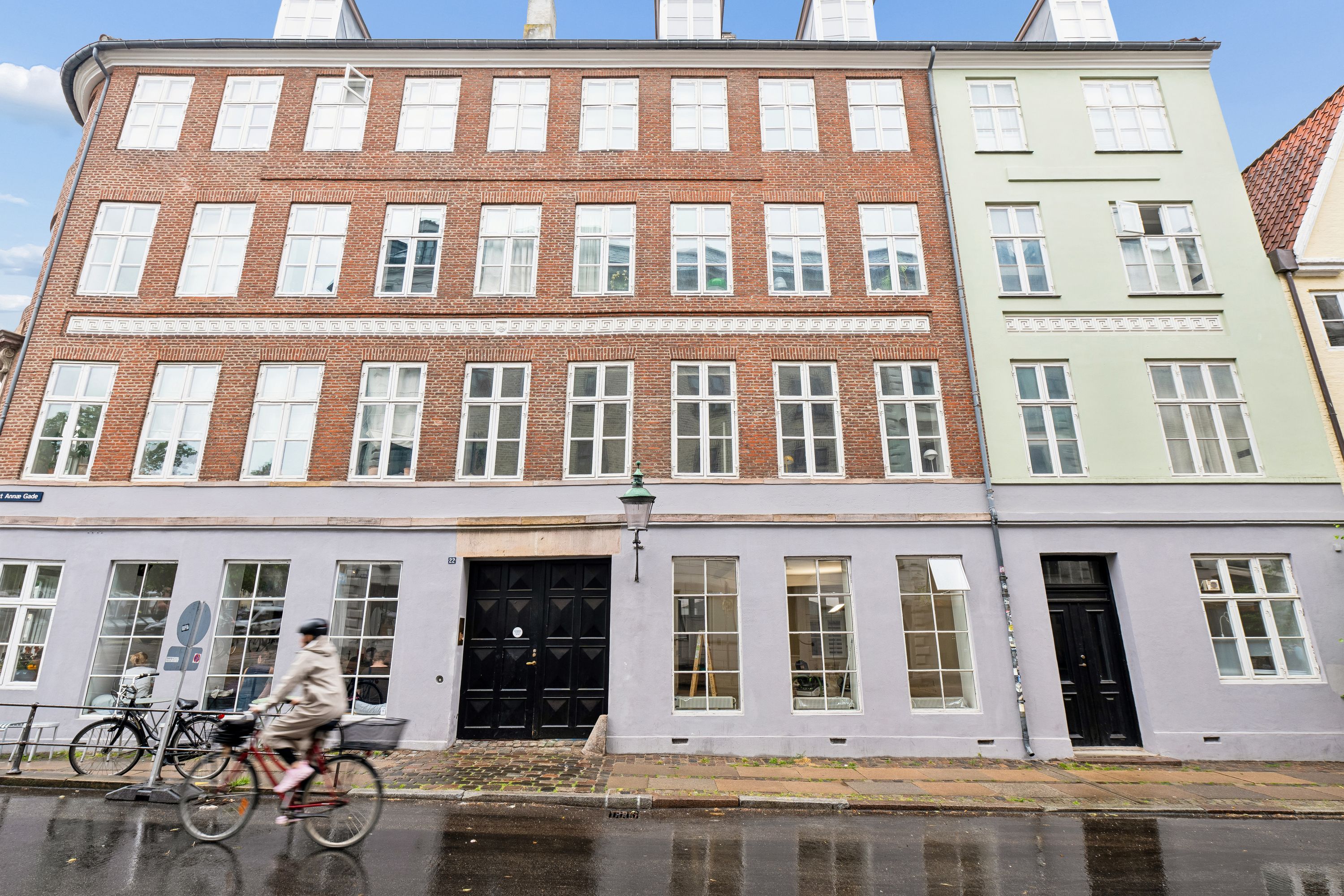 En cykelkører kører forbi en kontorbygning med røde mursten og store ruder, Sankt Annæ Gade 22, København. Bygningen har store sort-hvide døre og en gadelampe