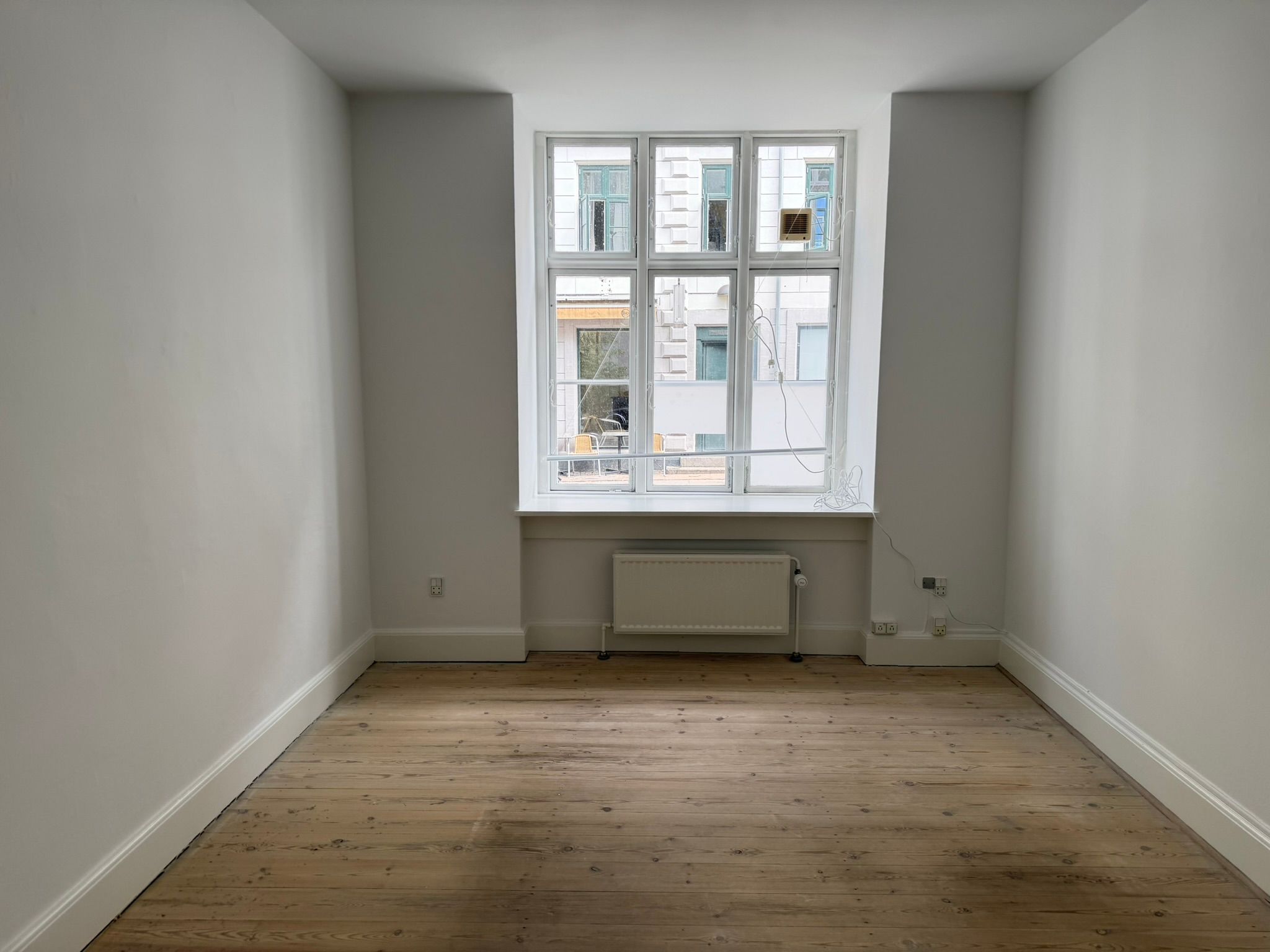 Ledig kontorrum med højloft, massivt trægulv, hvide vægge, radiator og tre store vinduer med udsigt til gaden med bygninger og terrasser