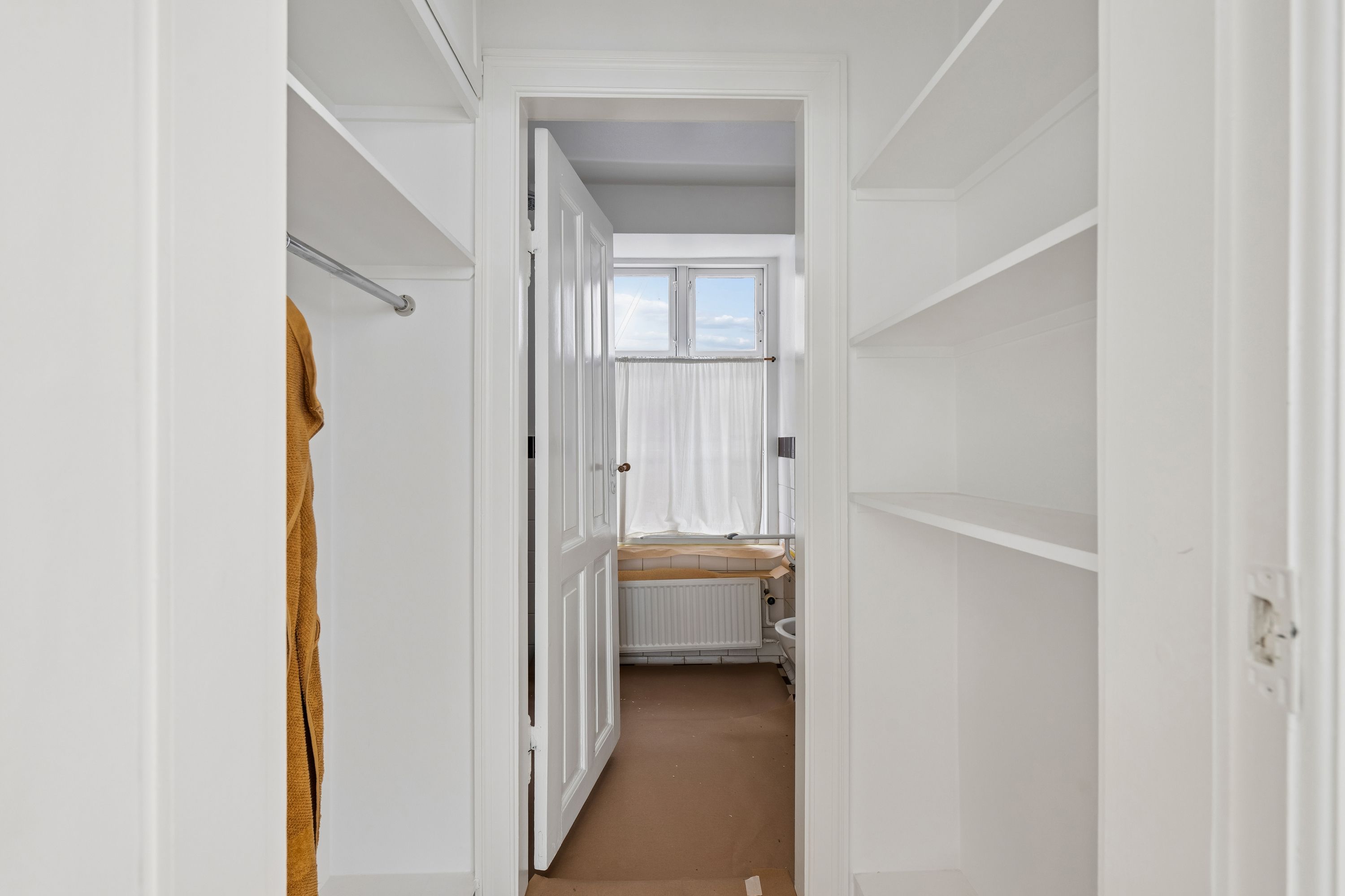 Kort korridor med lyse hvide vægge og højt til loftet. Dobbeltskabsgarderobe og to store hvidt hyldesystem med åbne døre. Dør med håndtag og hvid gardin til badeværelse med radiator og toilet.