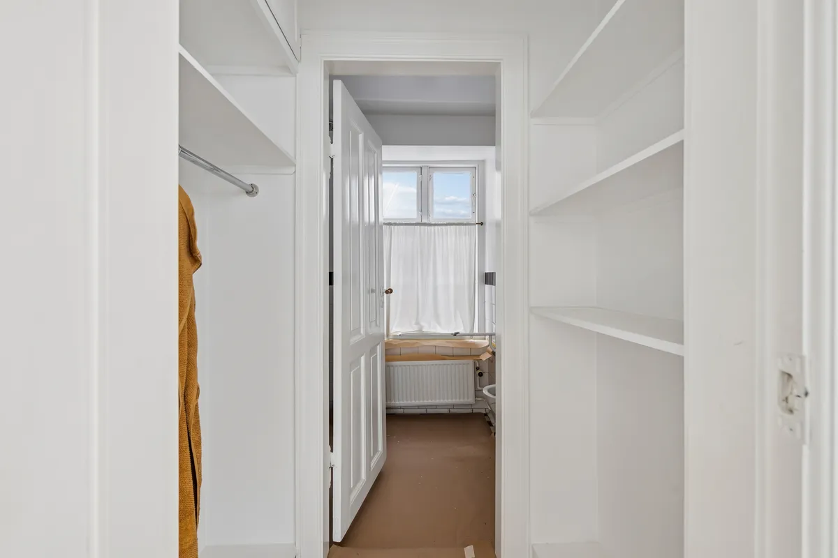 Kort korridor med lyse hvide vægge og højt til loftet. Dobbeltskabsgarderobe og to store hvidt hyldesystem med åbne døre. Dør med håndtag og hvid gardin til badeværelse med radiator og toilet.