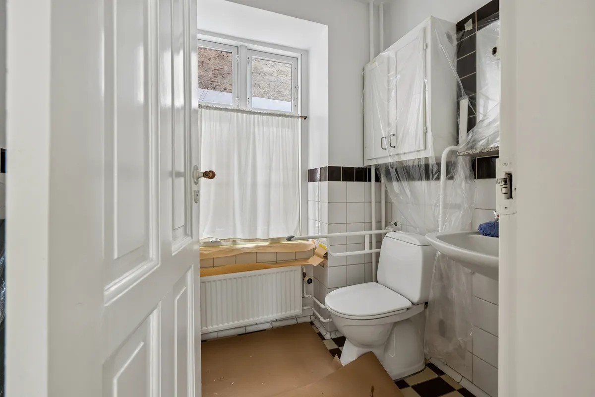 Et renoveret badeværelse med hvidt toilet, hvidt badekar, hvidt badeværelsesskab og hvidt badeværelsesvindue med hvid gardin og plastfolie på badeværelsesvæggene