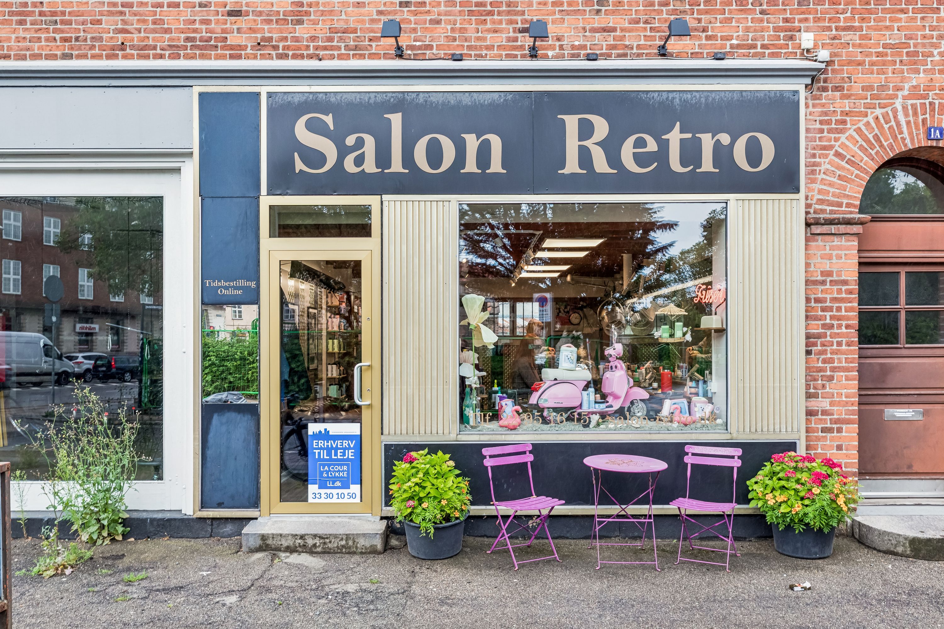 Lejlighedsvis butik til leje med terrassebord og stole og lyserøde planter og Salon Retro skilt på facaden.
