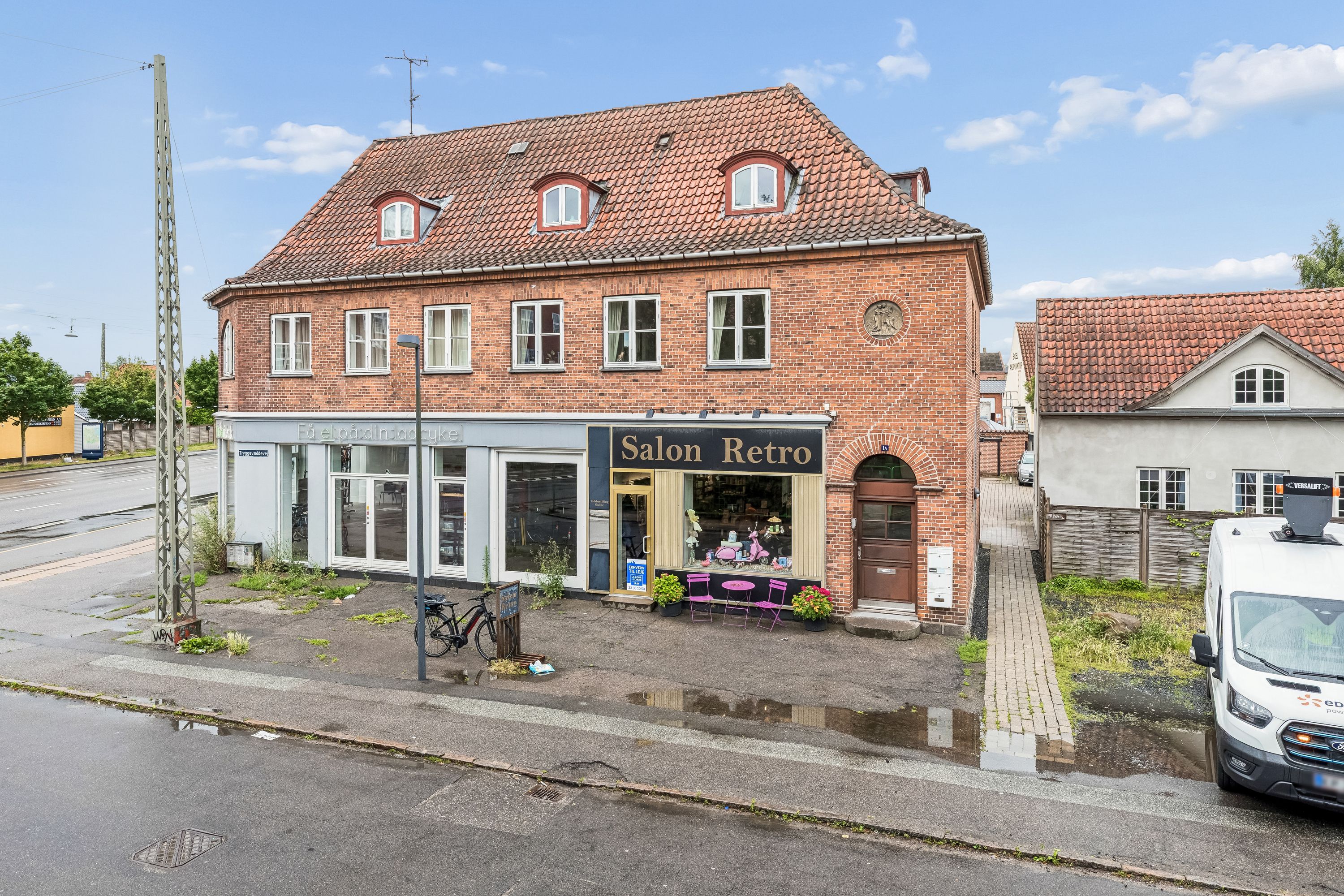 Butikslokaler til leje på Tryggevældevej 1A i Vanløse med salon og blomster på vej, hvor en cykel og en hvid varevogn er parkeret.