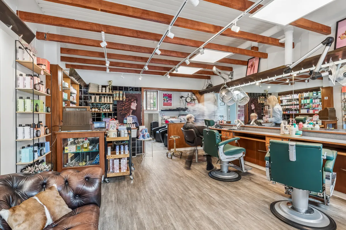 En barbersalon med lyse gulvbelægning, stole, tøjskabe, stole og lysekroner på loftet samt en sofa med puder i den ene ende