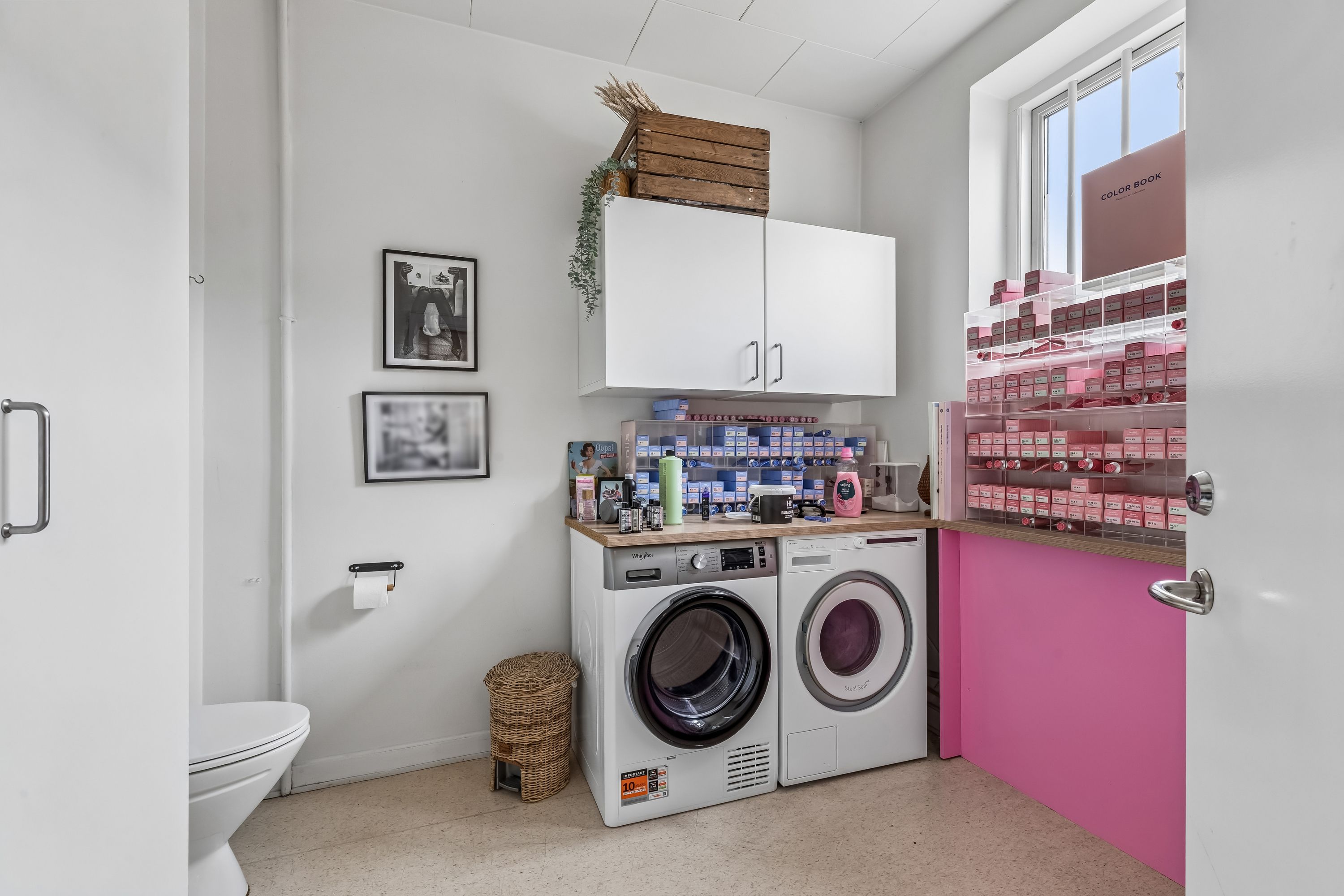 Hvid toilet med papirrullestativ, hvide kabinetter og to hvide tøjskabsmaskiner, en med pink farvede hylder og produkter, og to billeder på væggen, et med tekst og det andet med et menneske