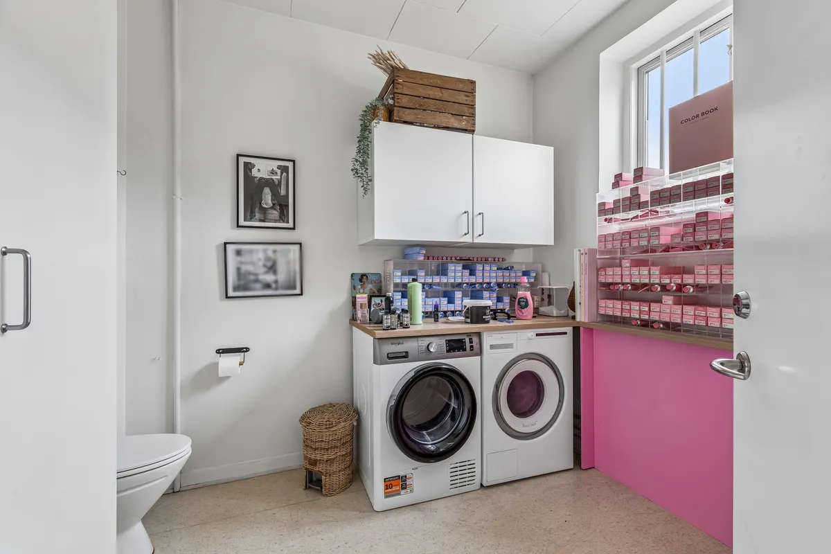 Hvid toilet med papirrullestativ, hvide kabinetter og to hvide tøjskabsmaskiner, en med pink farvede hylder og produkter, og to billeder på væggen, et med tekst og det andet med et menneske