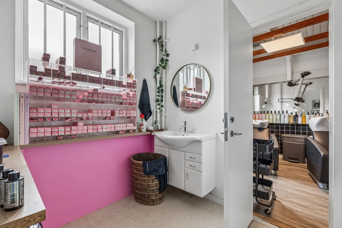Butikslokaler med højt til loftet, lyst og rummeligt med pink og hvide vægge, en stor spejl og et nyt toilet. I vinduet står et farvebog med røde bokse.