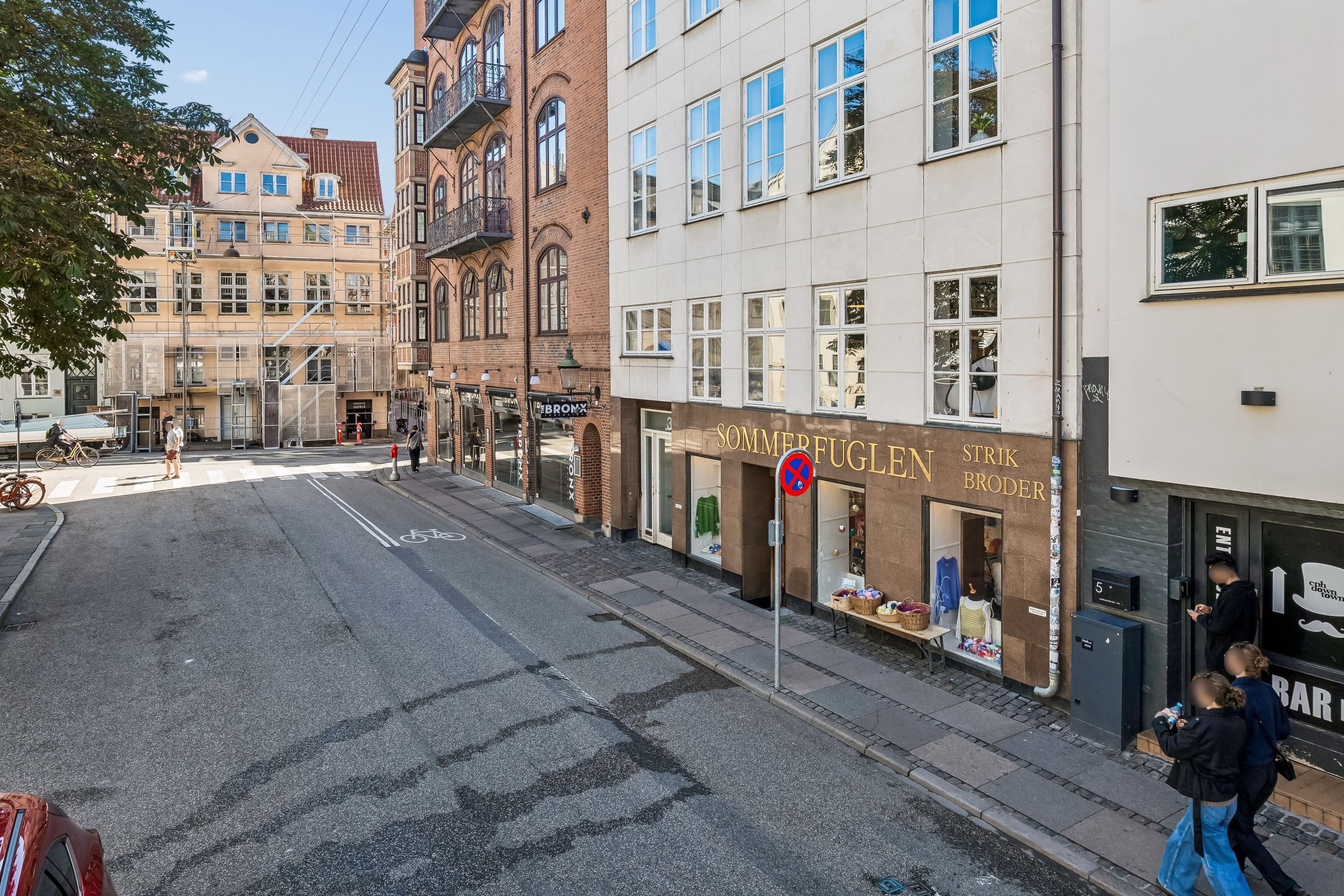 Kontorbyggeri med flere indgange og gadestegn. To mennesker går forbi et butiksvindue med teksten Sommerfuglen. En mand har en vandflaske og en kvinde har en taske
