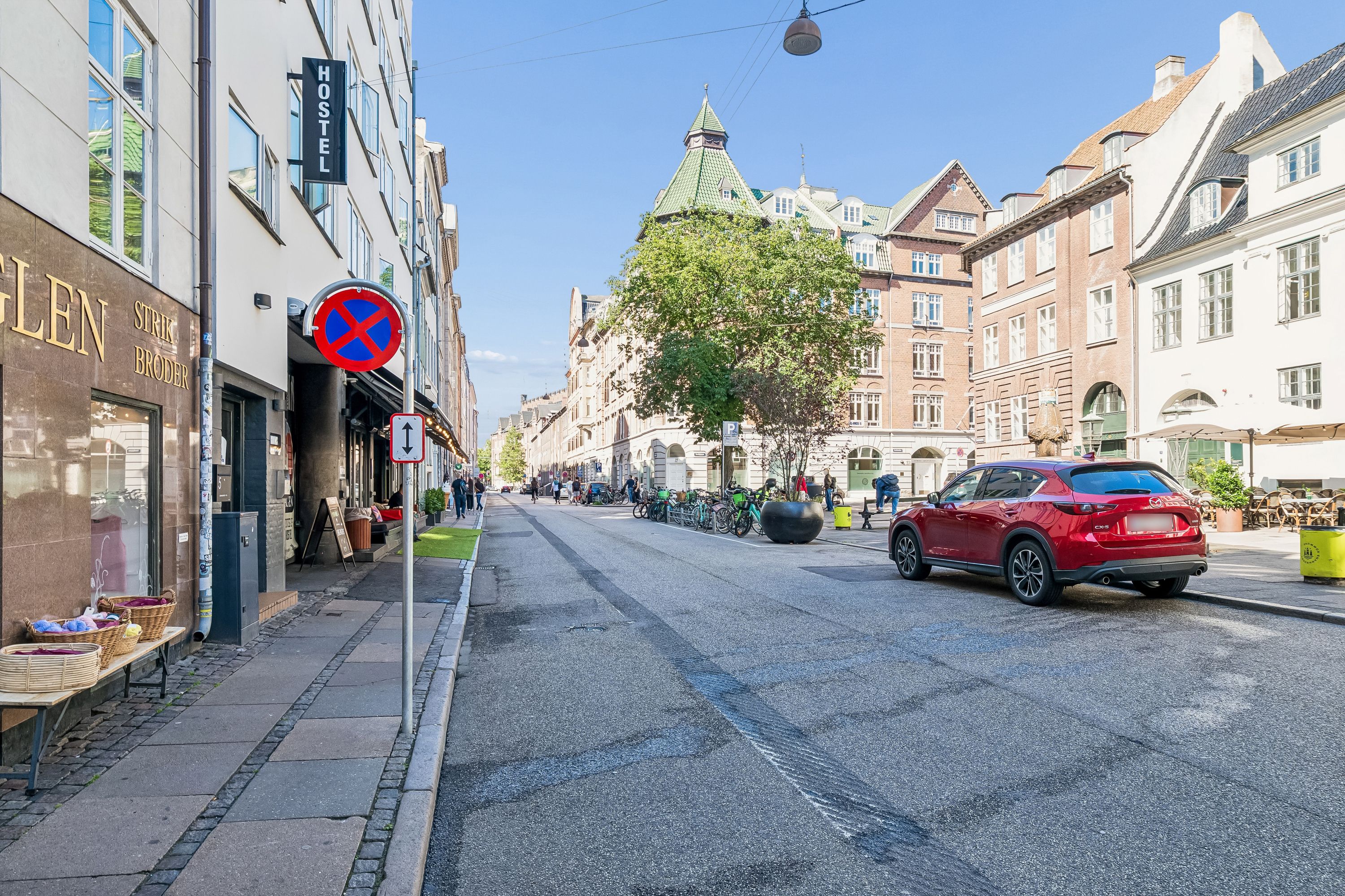 Rød crossover parkeret i gade, caféer, grønne træer og blomster, gul skrift Hostel, lyseblå himmel, vejledningsskilt og gavlede bygninger i baggrunden