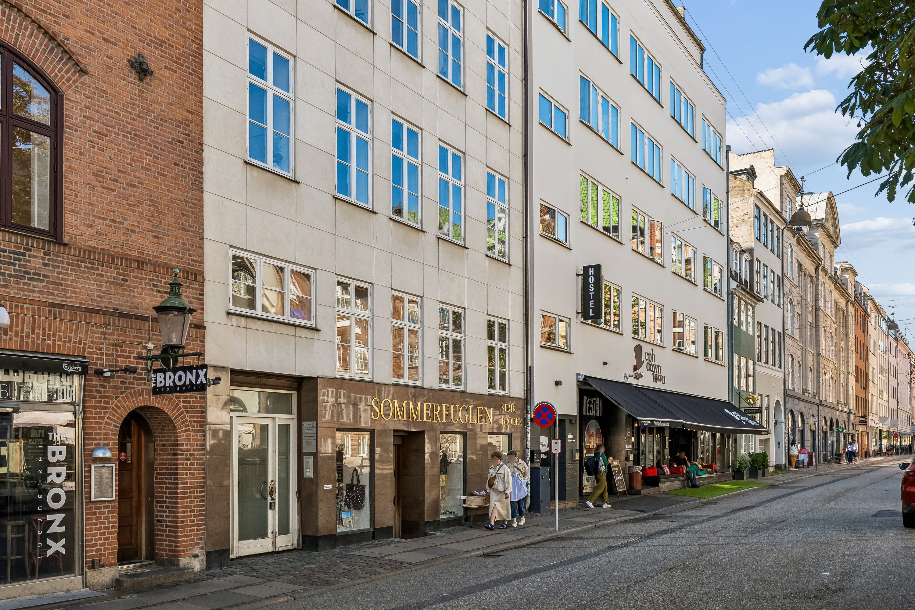 Kontor til leje på Vandkunsten 3 med lyse vinduer og elegant facade. En kvinde og mand går forbi et butiksvindue med teksten Sommerfuglen. En rødvognen er parkeret ved siden af gaden
