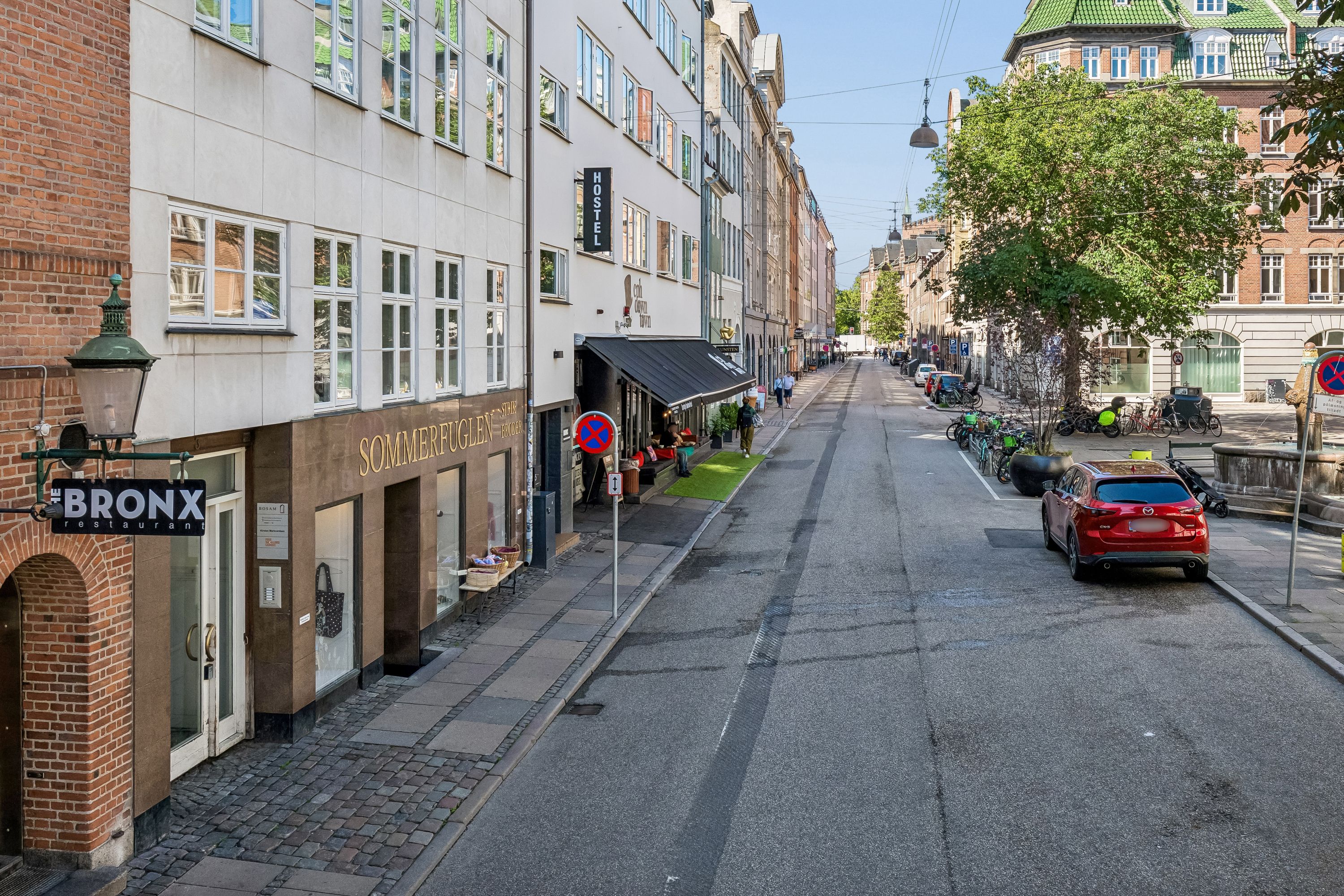 Gåsetraede gadeskæg med et parkerede bil og cykler og grønne buske og stående lygter langs fortovet og røde skilte med mærket Bronx og Sommerfuglen