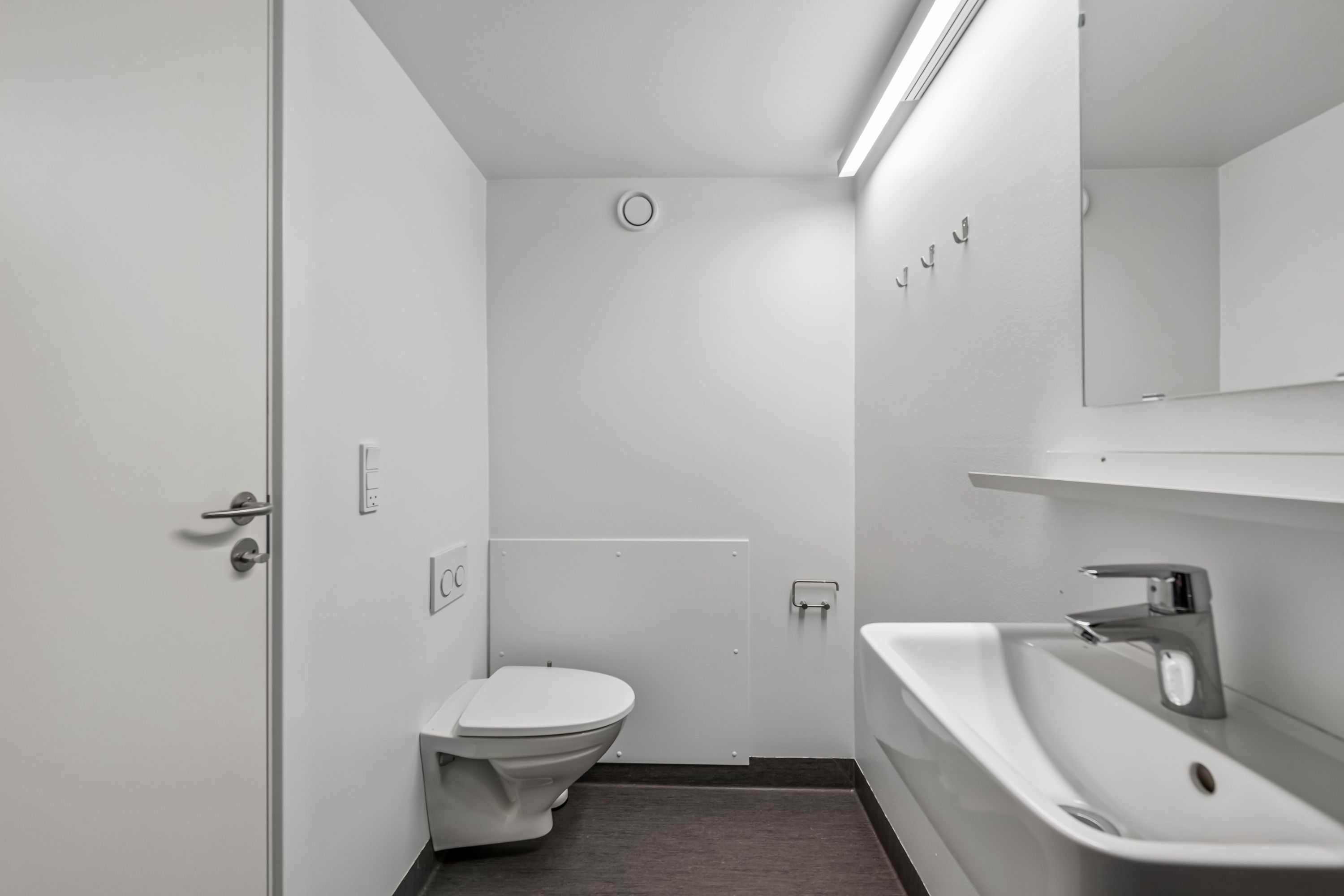 En lukket dør med håndtag og to nøglehuse og et toilet med toiletpapir i en hvid toiletrum med spejl og hængehylde