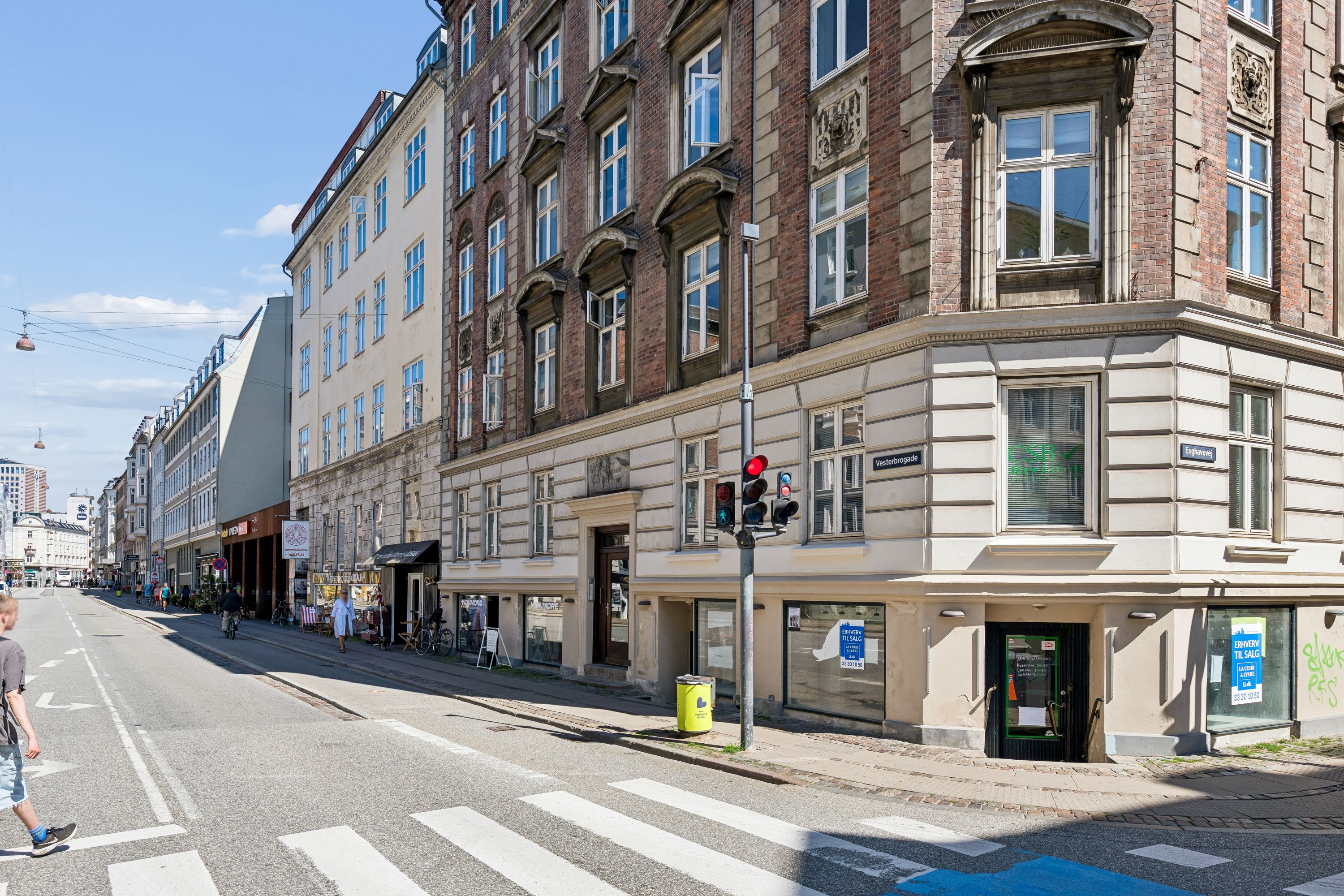 Et stort erhvervslokal med flere vinduer og to indgange på Vesterbrogade. Folk cykler langs gaden og krydser en zebraovergang.