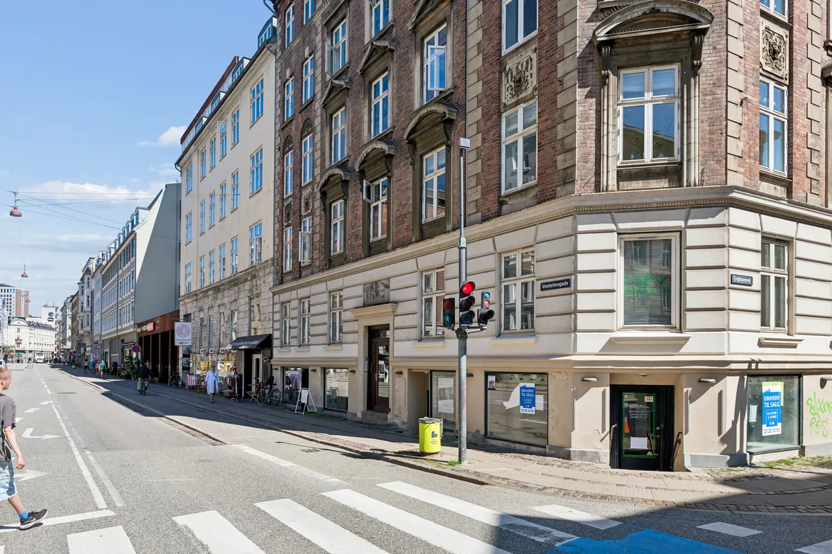 Et stort erhvervslokal med flere vinduer og to indgange på Vesterbrogade. Folk cykler langs gaden og krydser en zebraovergang.