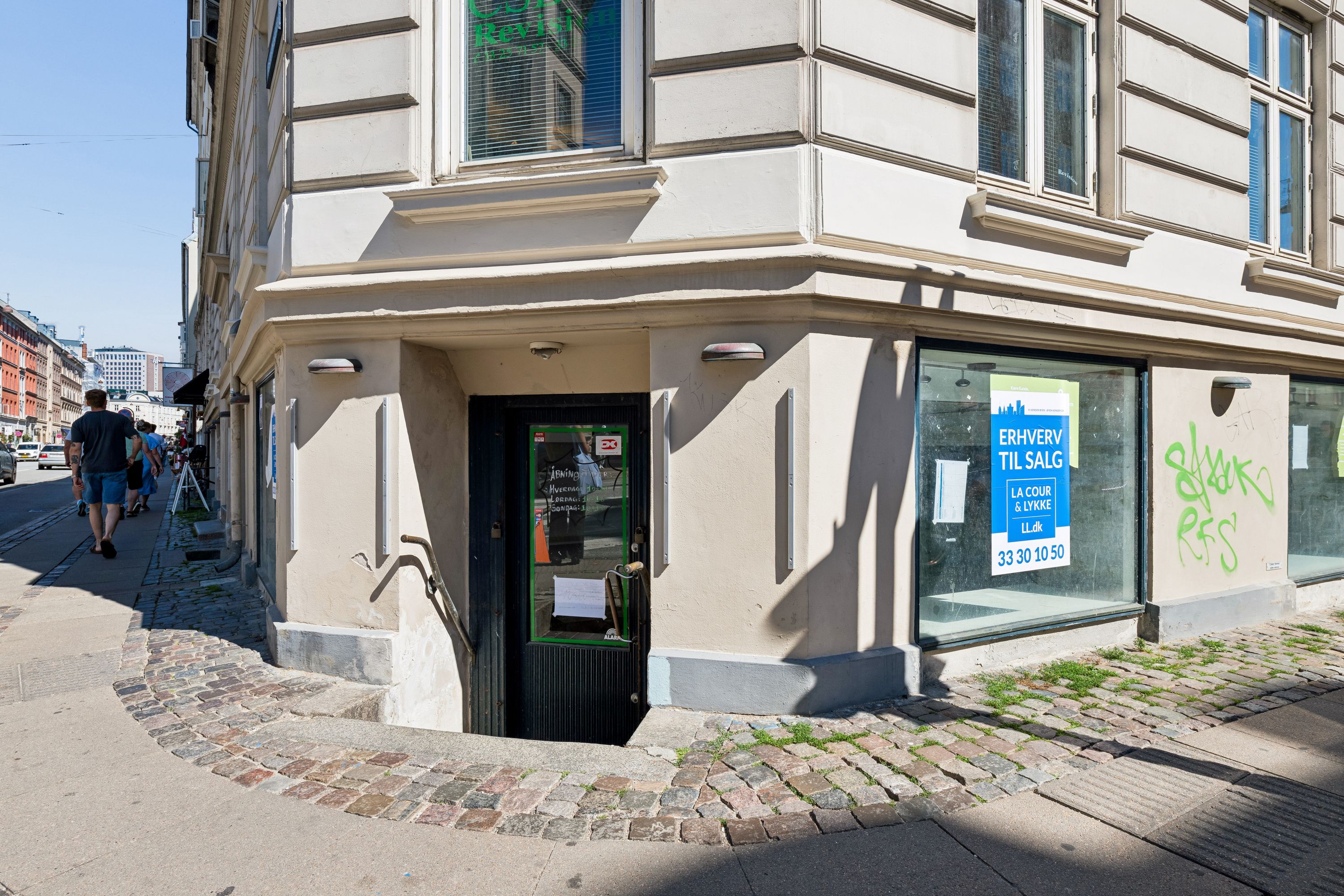 På Vesterbrogade 101 står et erhvervslokal med glasvinduer og skilte, der viser salg. Bygningen har gulmalede vægge og erhvervslokalet er i kælderplan. To voksne går langs fortovet og skyggerne falder på gaden