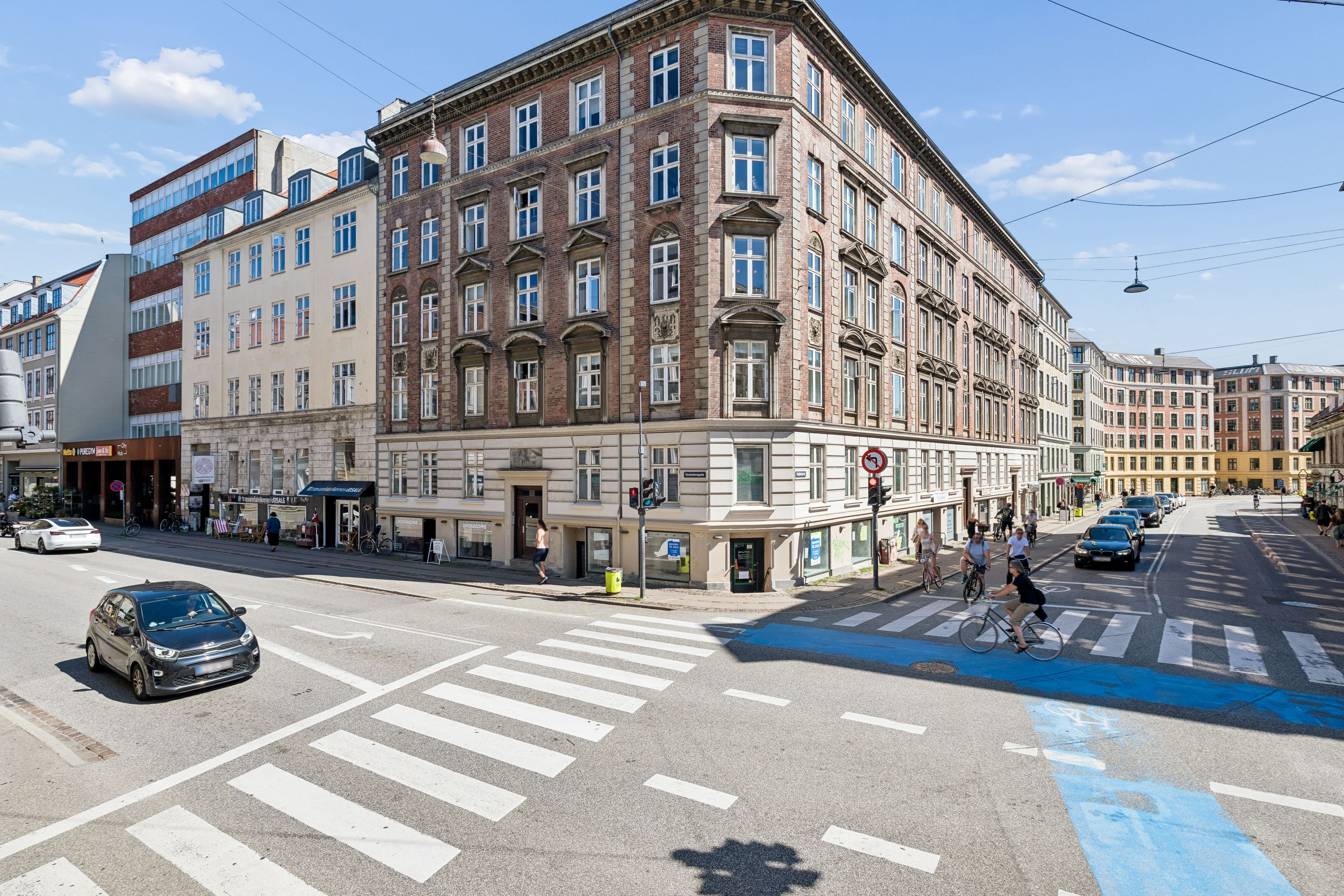 Forretningslokaler til salg på Vesterbrogade i København. Folk går og cykler i centrum. Biler og cykler er parkeret langs gaden. Bygningen har flere etaper og vinduer