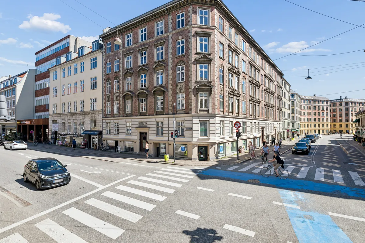 Forretningslokaler til salg på Vesterbrogade i København. Folk går og cykler i centrum. Biler og cykler er parkeret langs gaden. Bygningen har flere etaper og vinduer