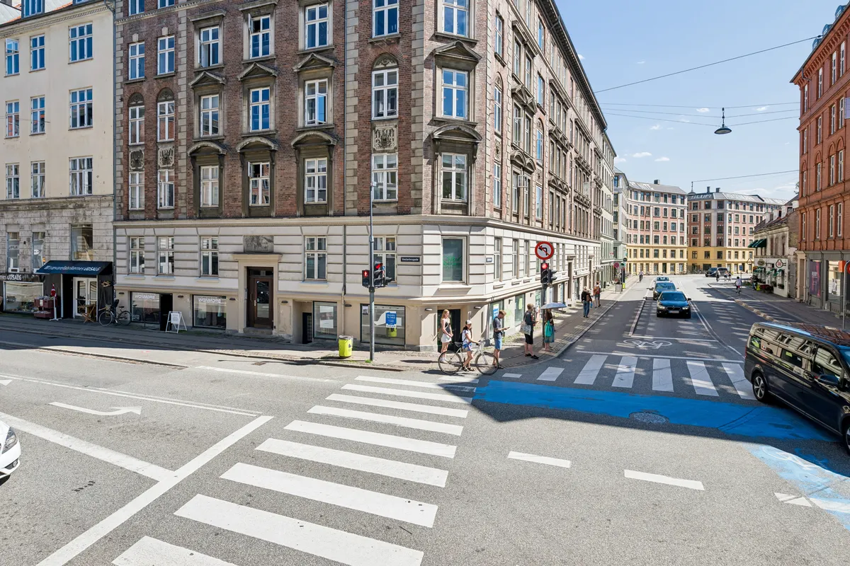 Flere biler og en minibus kører ad Vesterbrogade, hvor mange mennesker krydser fodgængerovergangen og går forbi forskellige butikker og huse med mange vinduer