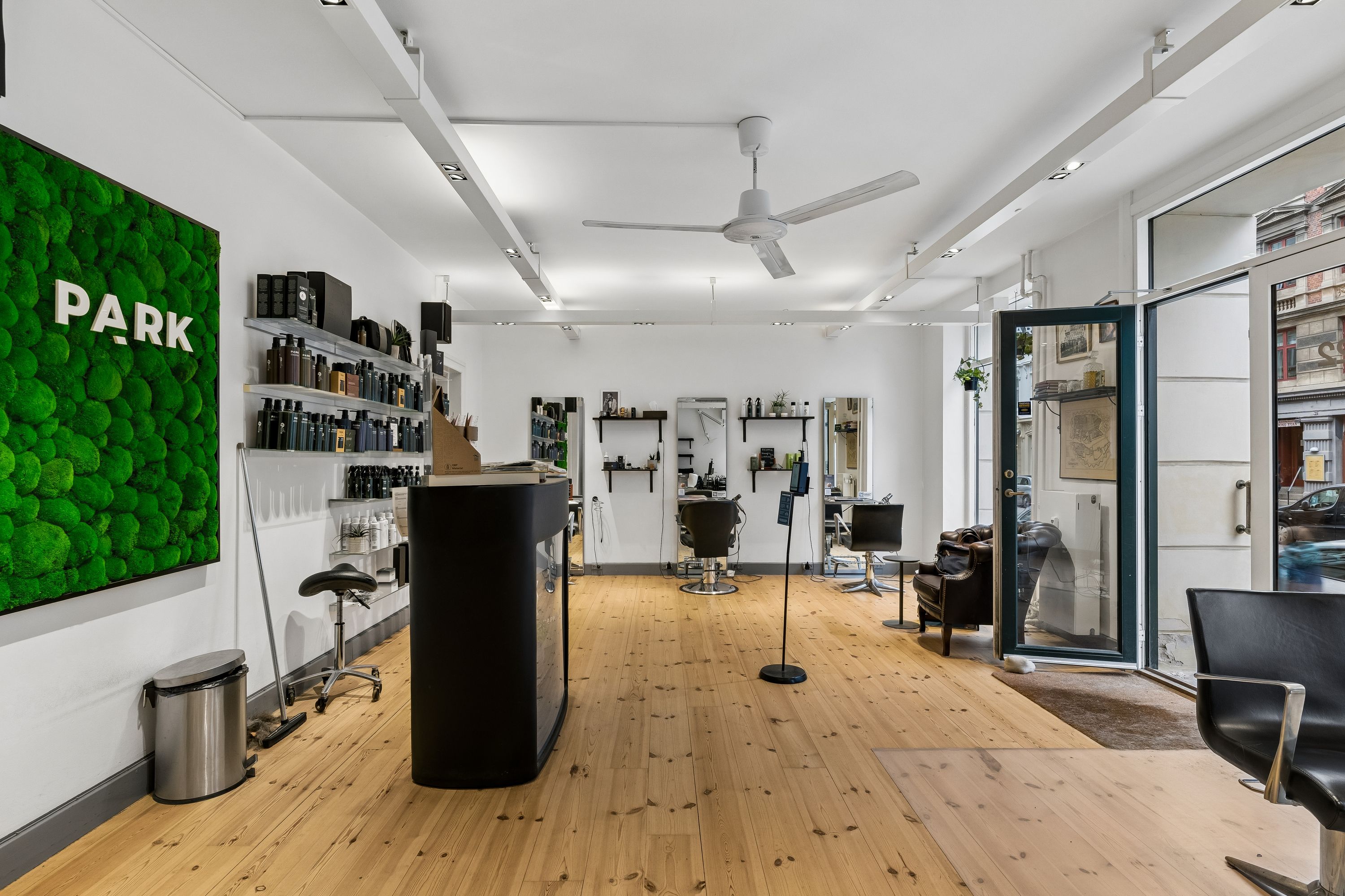 Inde i frisørsalonen ser man moderne møbler, en hvid væg og et grønt vægmaleri med ordet Park. Der er stolpeventilatorer og glasdøre.
