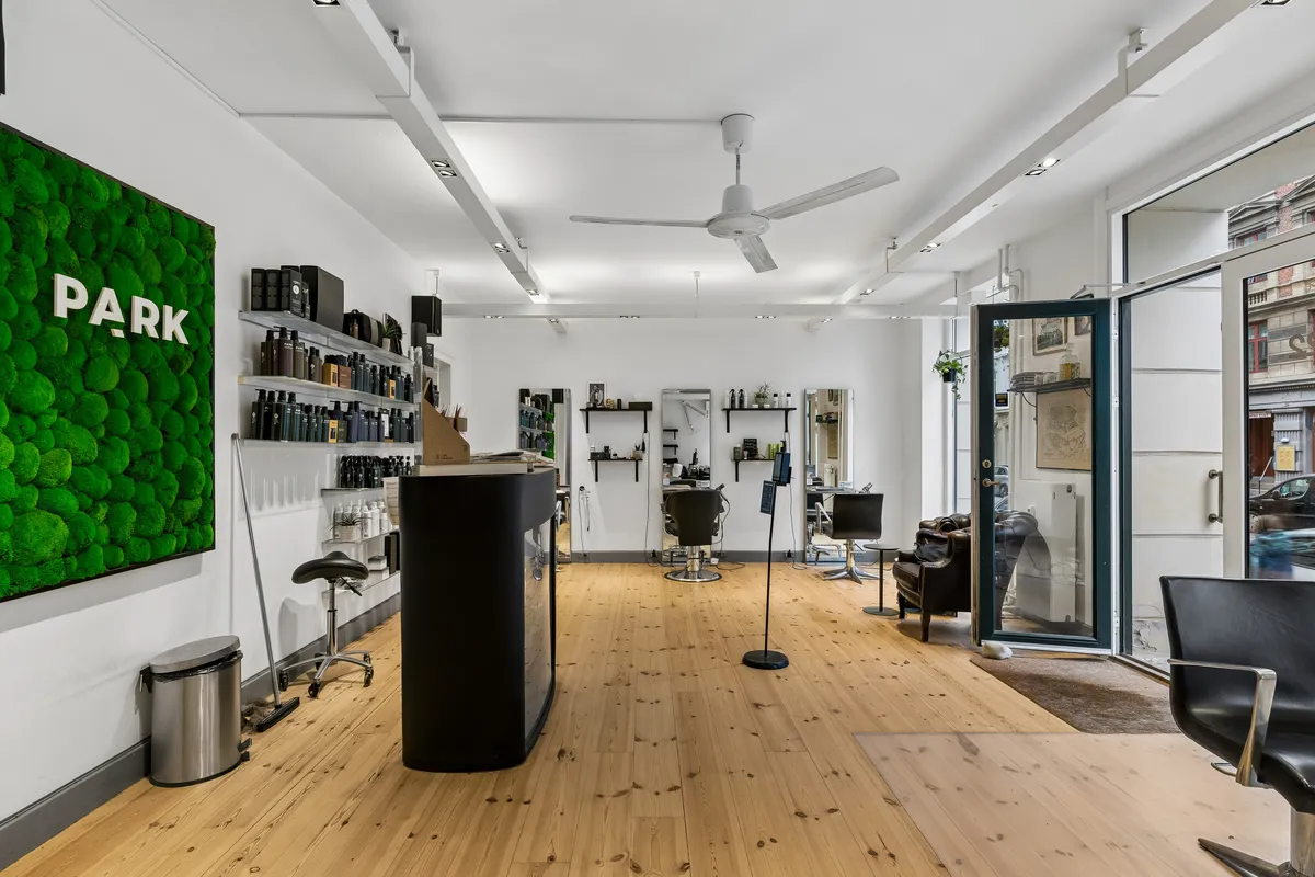 Inde i frisørsalonen ser man moderne møbler, en hvid væg og et grønt vægmaleri med ordet Park. Der er stolpeventilatorer og glasdøre.